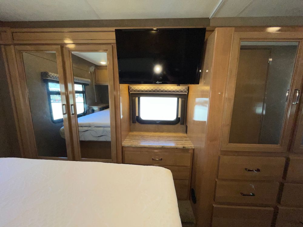 RV rental interior: bedroom features bed with white linens, wall-mounted TV, mirrored wardrobe, and dresser with window.. Thor Quantum 2019