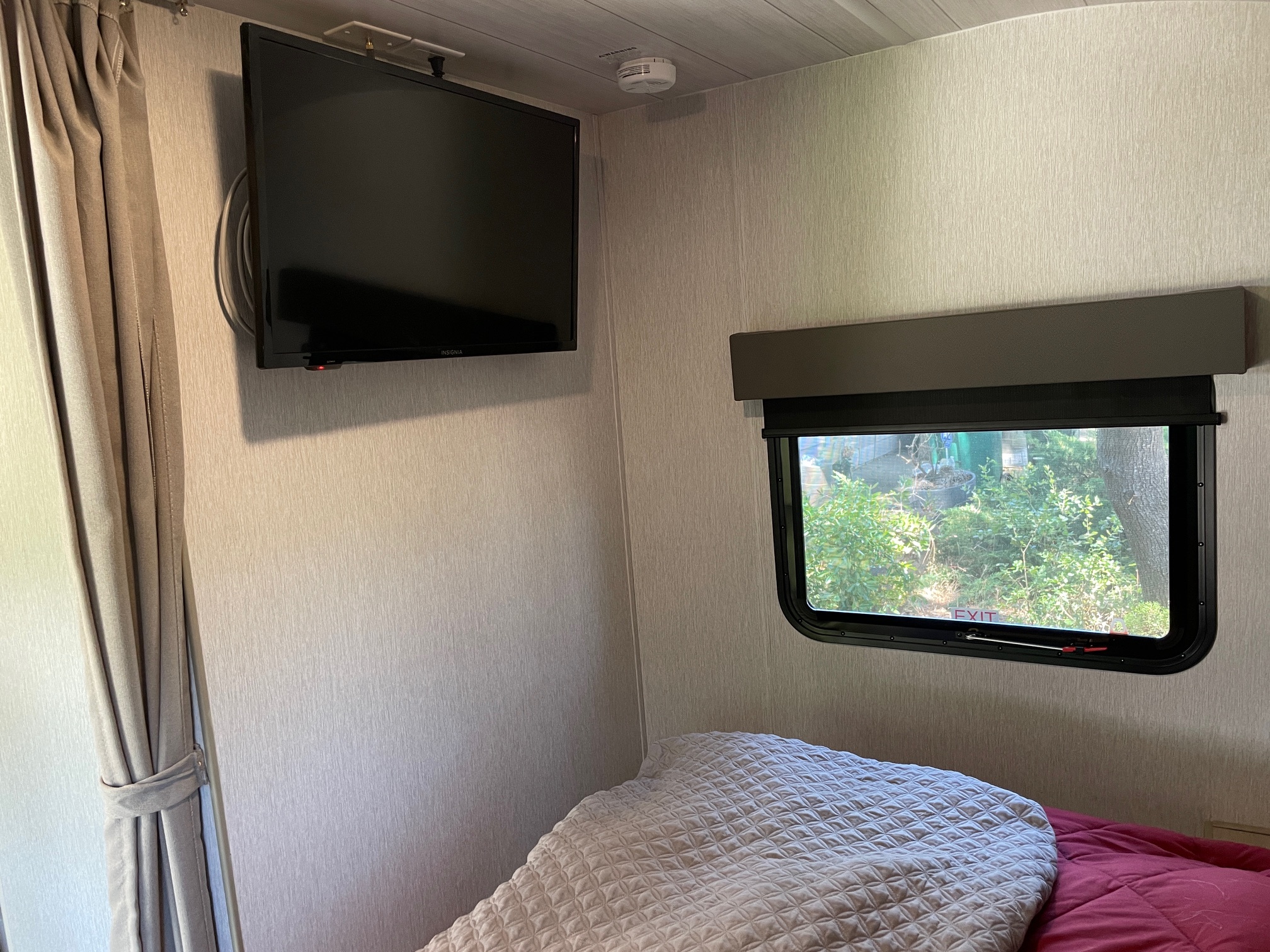Compact RV rental bedroom featuring a wall-mounted TV, window with green view, and cozy bed.. Forest River Wildwood X-Lite 2023