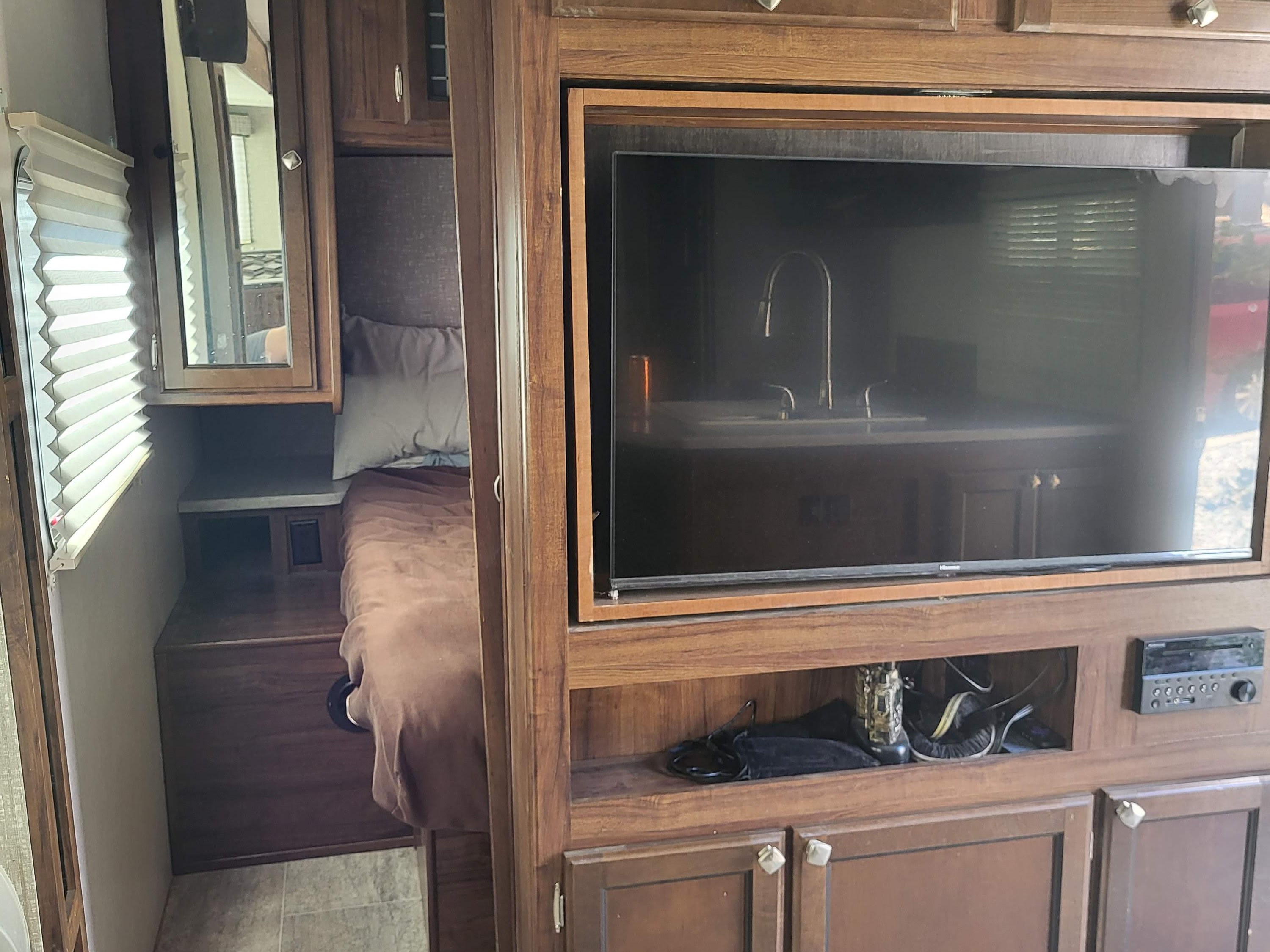 Cozy RV rental interior shows a compact bed, window with blinds, and TV/stereo setup, reflecting a kitchen area.. Heartland RVs Mallard 2018