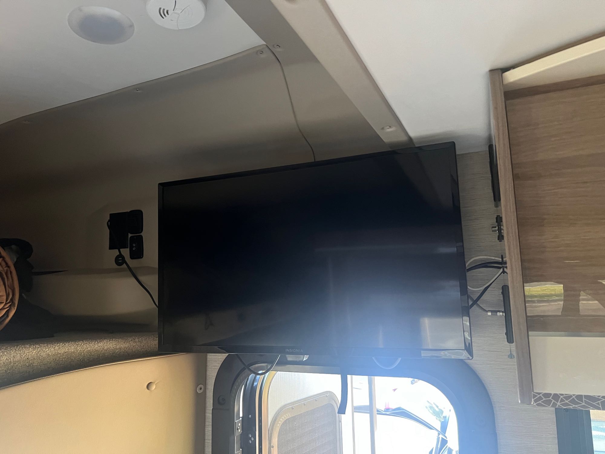 RV rental interior: Wall-mounted TV in sleeping area, with window below. Ceiling speaker & smoke detector visible.. Jayco Melbourne Prestige 2021