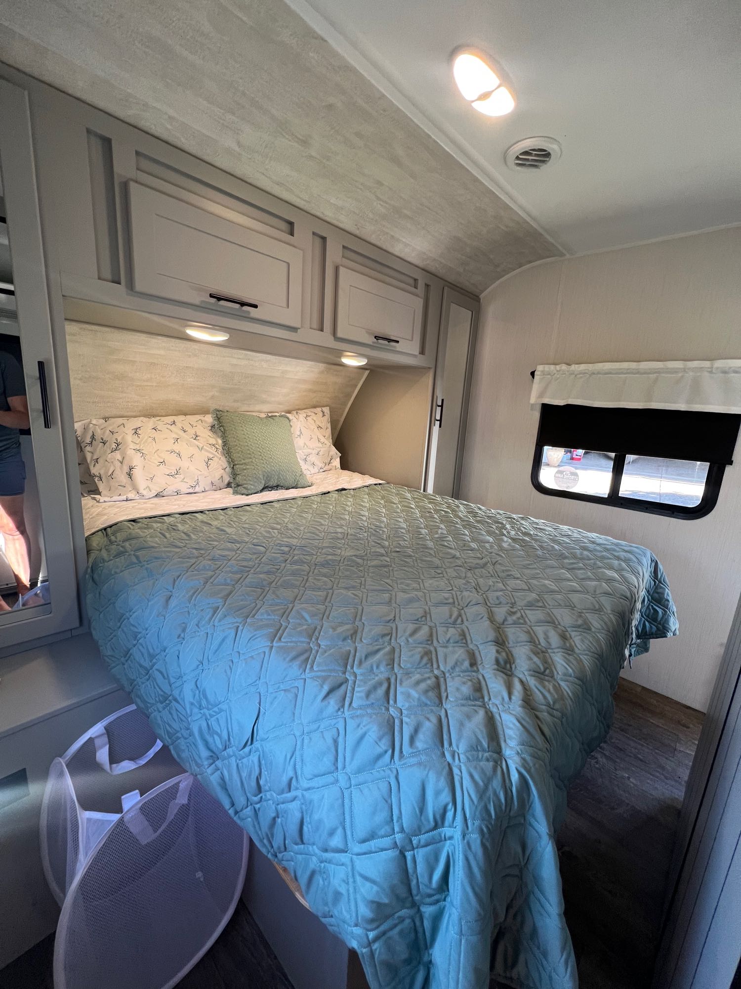 Experience comfort in this RV rental bedroom featuring a queen bed with blue quilt, overhead storage, and a window.. Forest River Rainier 24DBR 2022