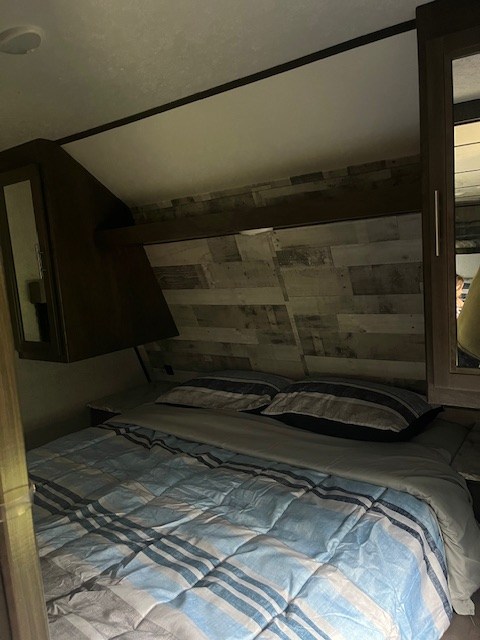 Cozy RV rental bedroom with blue striped bedding, overhead storage, and wood paneling above the bed.. Dutchmen Coleman Lantern 2020