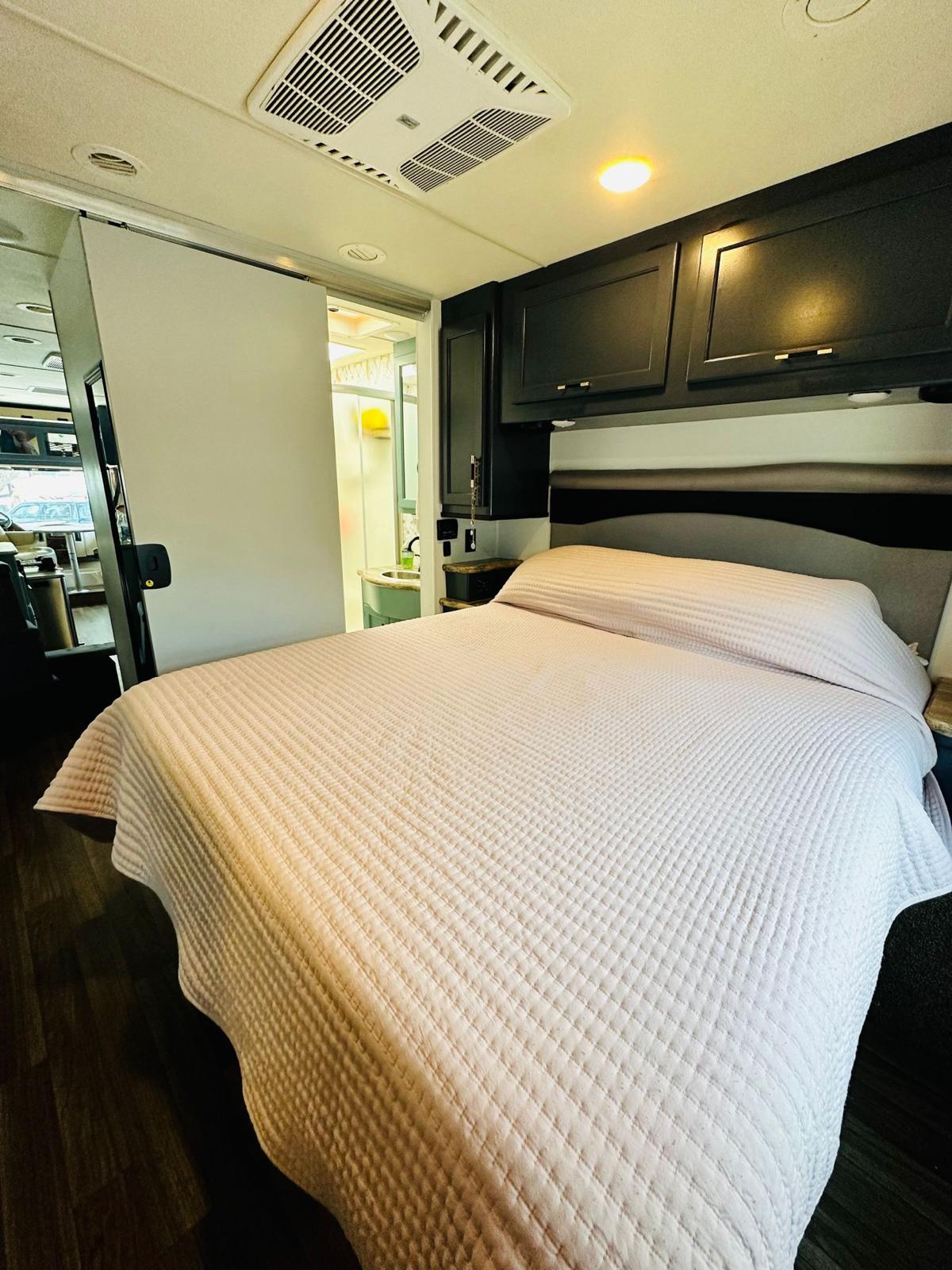 Inside an RV rental: a cozy bedroom with quilted bed, overhead cabinets, AC, and a sliding door to the bathroom.. Thor Motor Coach A.C.E 2018