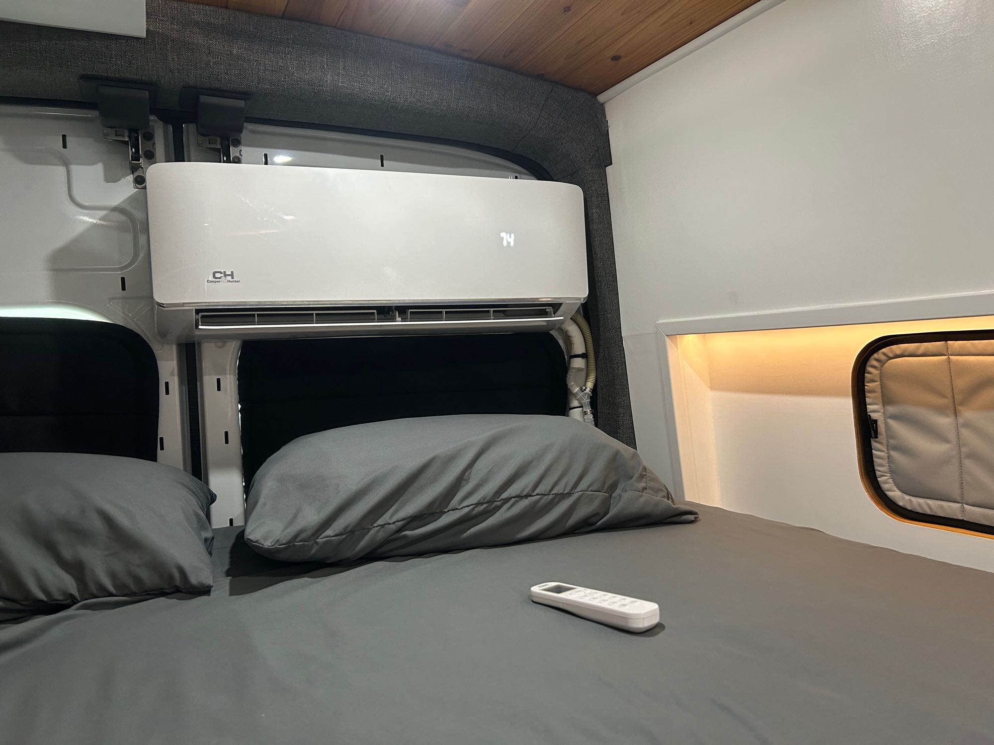 Inside an RV rental: a plush bed with grey linens, and an AC unit keeping the space cool at 74°F for a comfy journey.. Ford Transit 2018