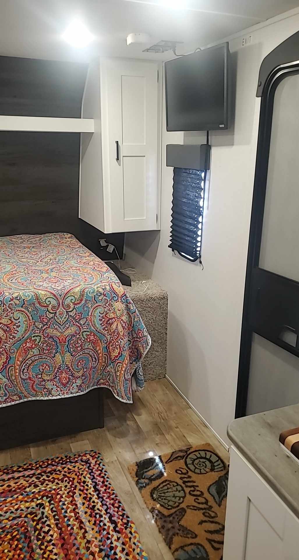 Cozy RV rental interior shows a colorful bed, white overhead cabinets, a wall-mounted TV, and inviting floor mats.. Keystone RV Hideout 2023