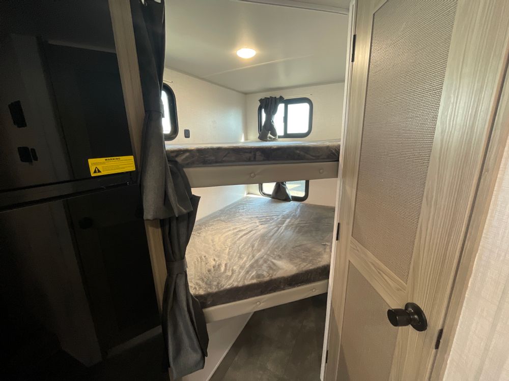 RV rental interior showcases stacked bunk beds with windows, a dark appliance, and a light wood door.. Forest River Wildwood 2024