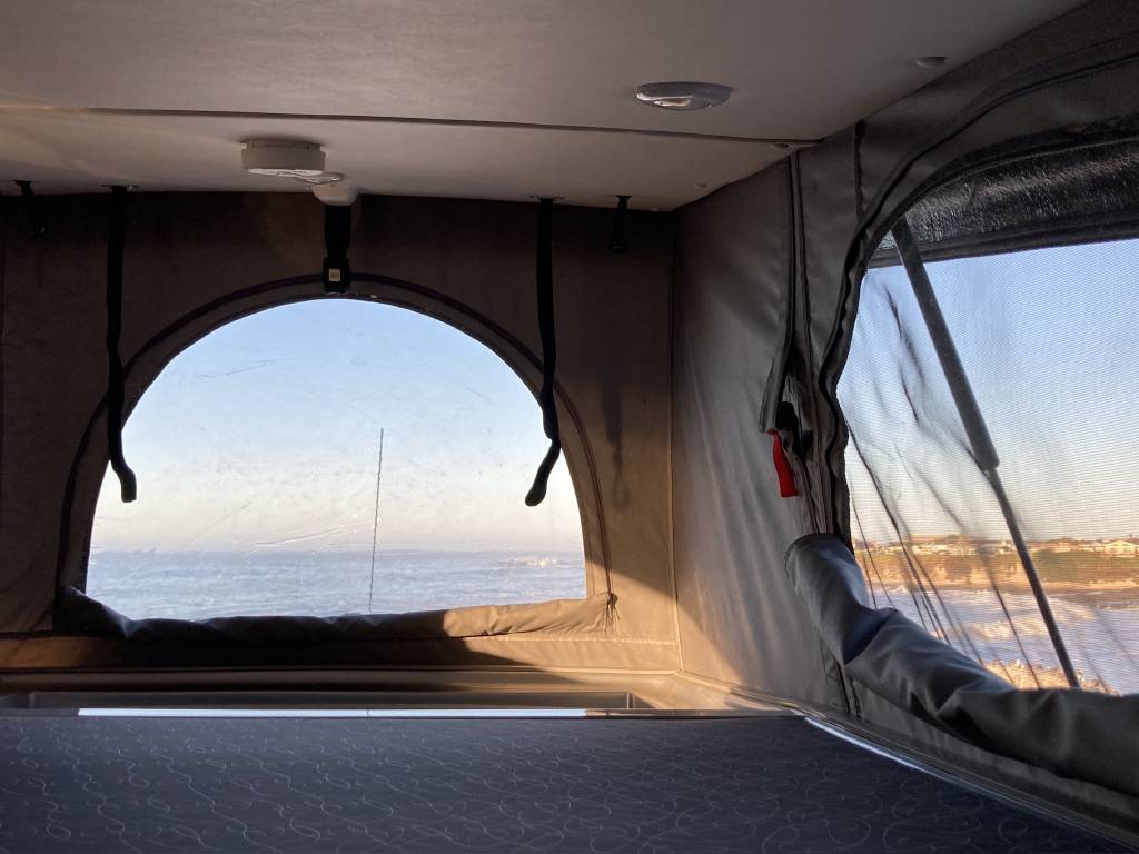 RV rental interior with stunning ocean views from pop-top clear and mesh windows. Includes a bed for coastal camping.. Pleasure-Way Tofino 2022