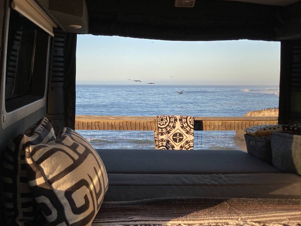 Ocean view from an RV rental: a cozy bed overlooking calm waters, flying birds, and distant waves. Van life travel.. Pleasure-Way Tofino 2022