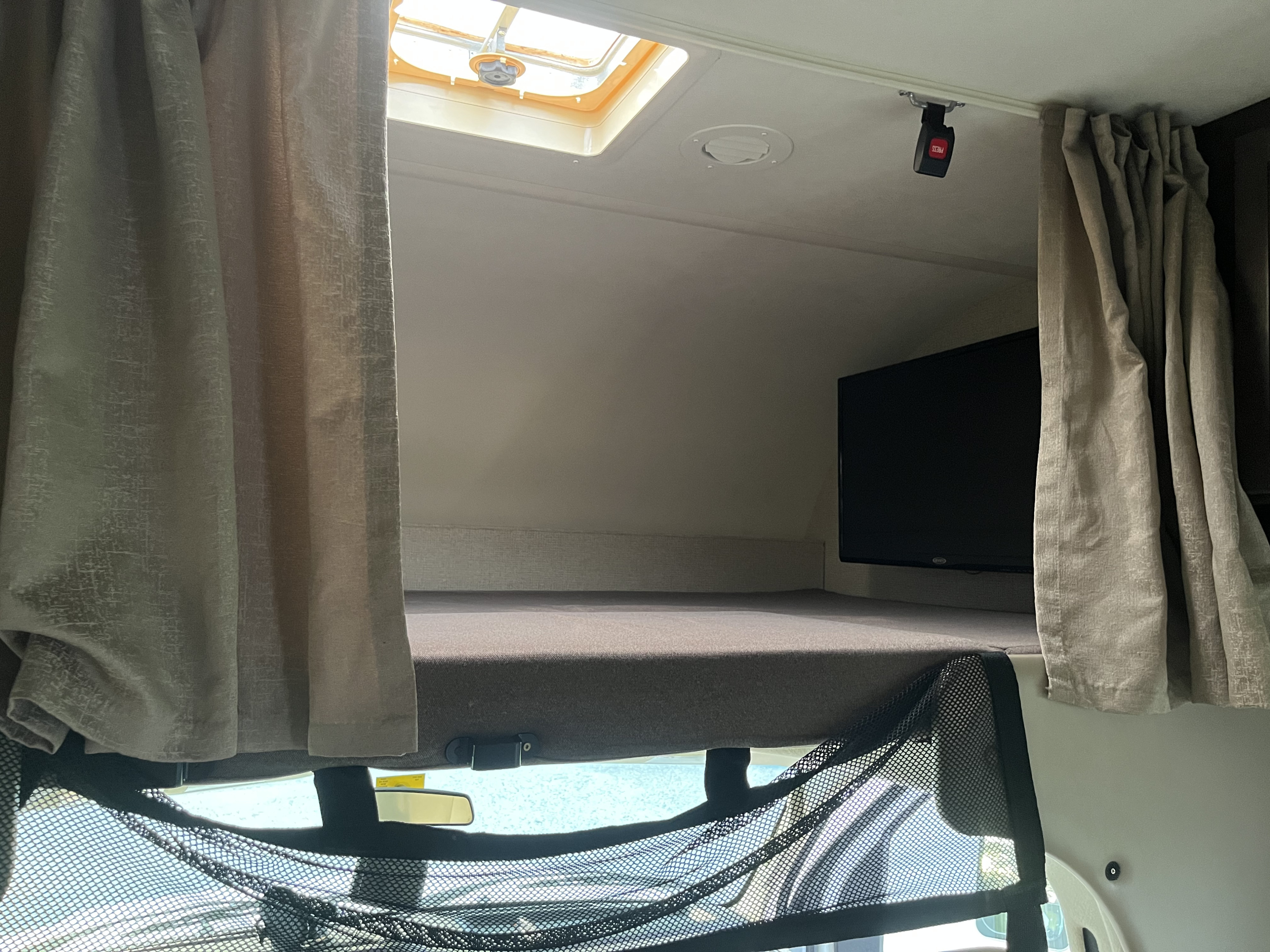 Overhead bunk area in an RV rental, showing a bed, mounted TV, skylight, and privacy curtains.. Thor Four Winds 2019