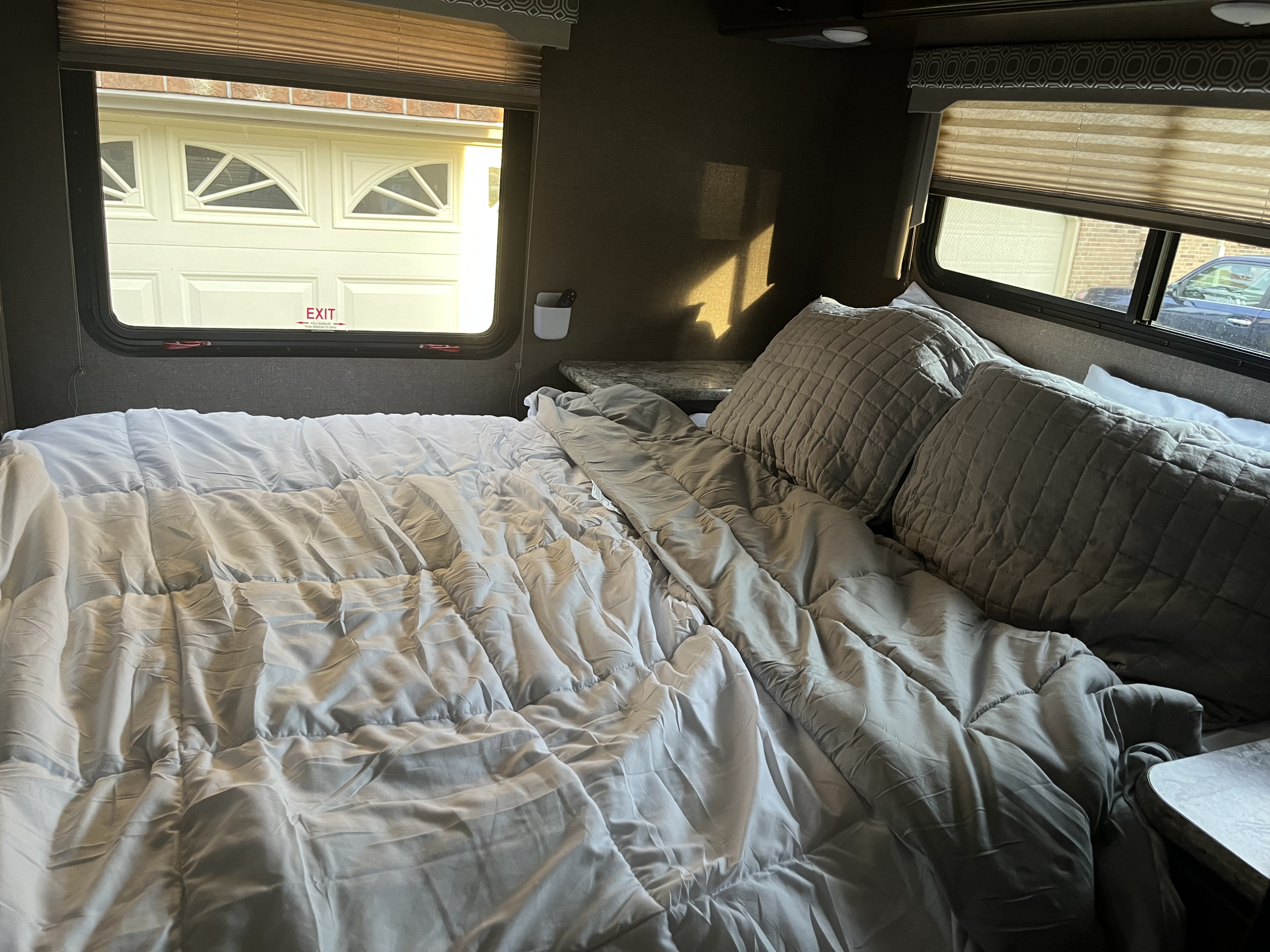 Cozy RV rental bedroom with a comfy bed, sunlit windows, and an "EXIT" sign, parked near a garage.. Thor Four Winds 2019