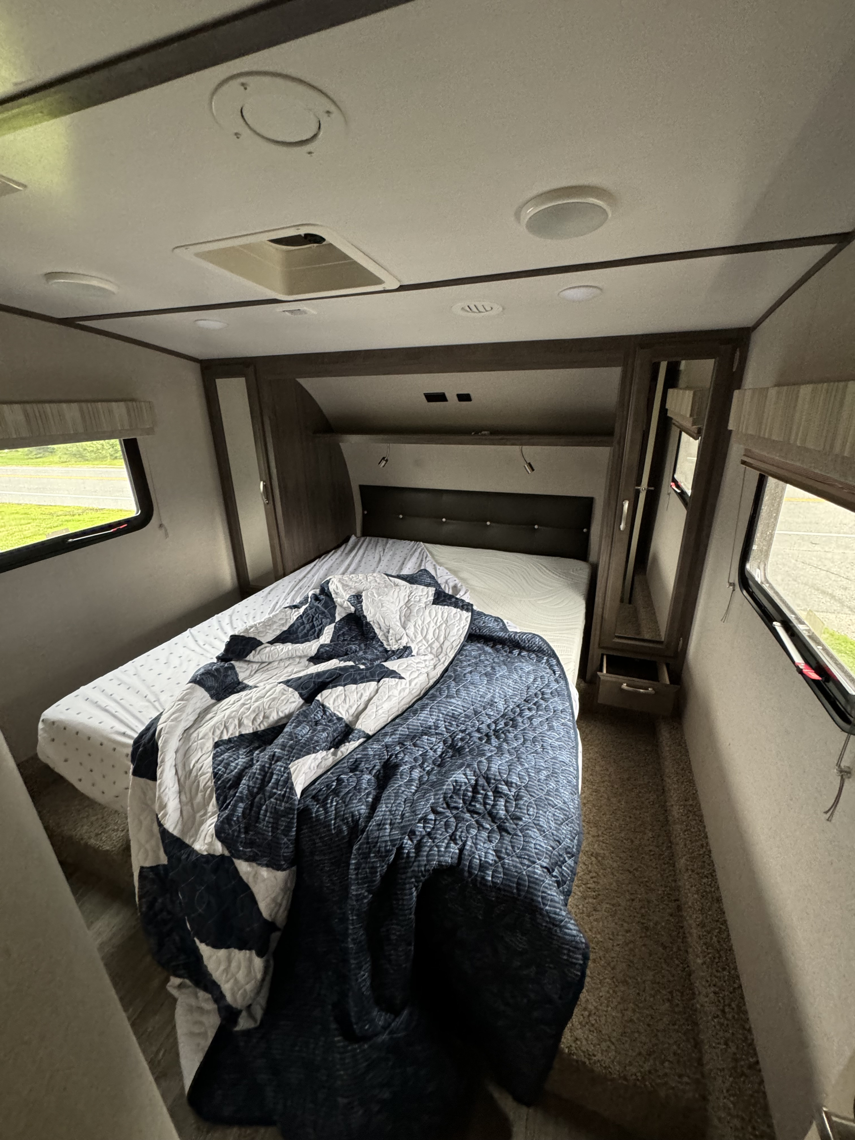 View inside an RV rental bedroom showing a bed with a blue and white quilt, a window overlooking a road, and ceiling fixtures.. Minnie Minnie Plus 2019