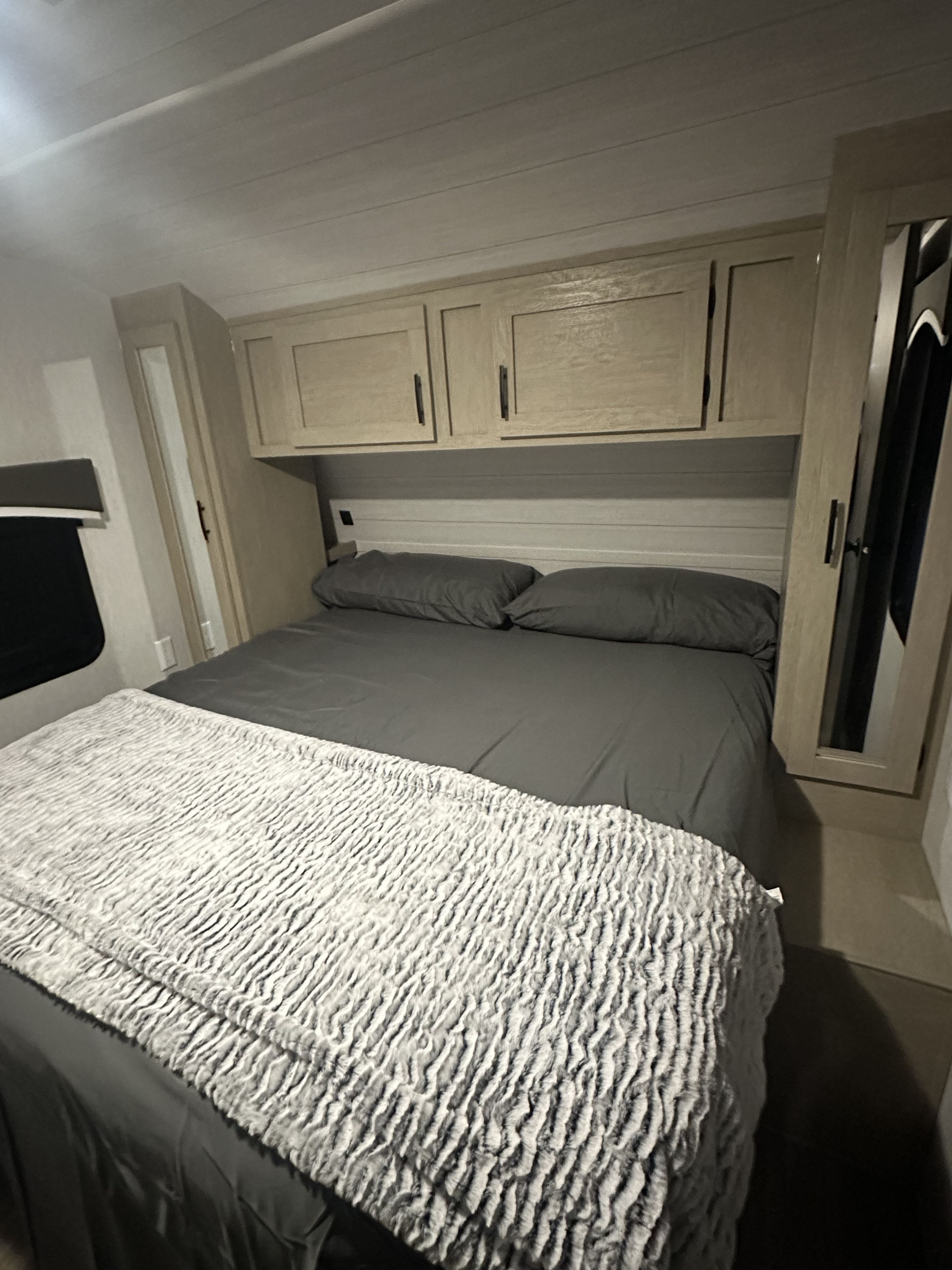 Cozy RV rental bedroom features a compact grey bed, overhead cabinets, and soft textured blanket for travel.. Forest River Evo 2023