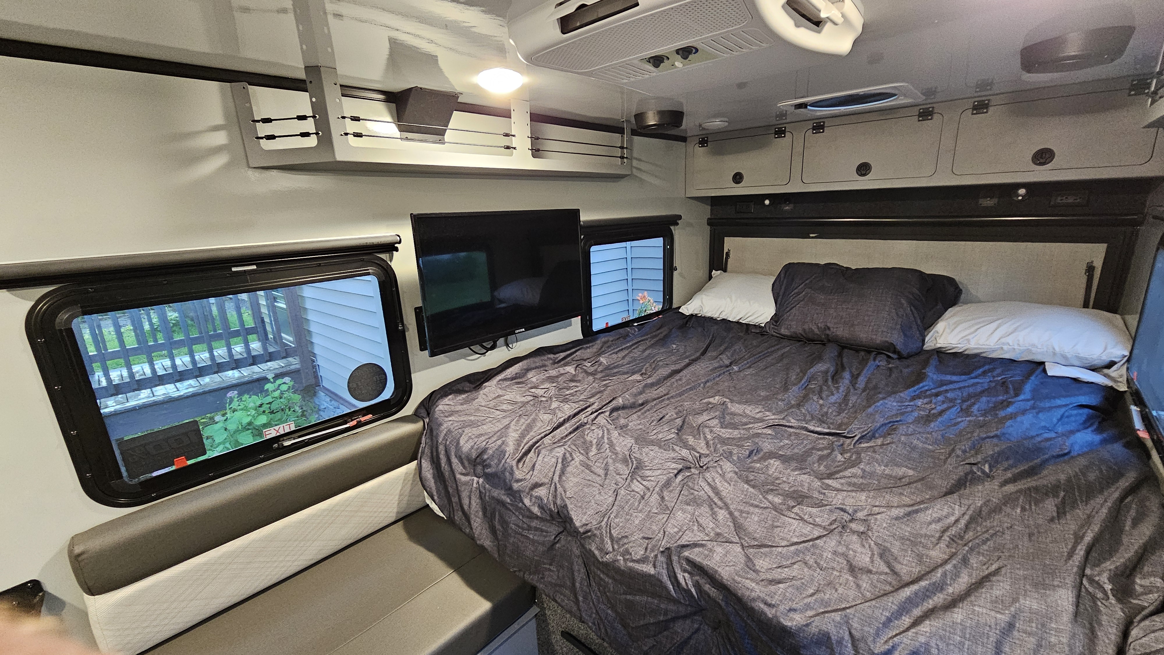 Inside a comfy RV rental bedroom: large bed, wall-mounted TV, windows, and overhead cabinets for storage.. Encore RV ROG 2023