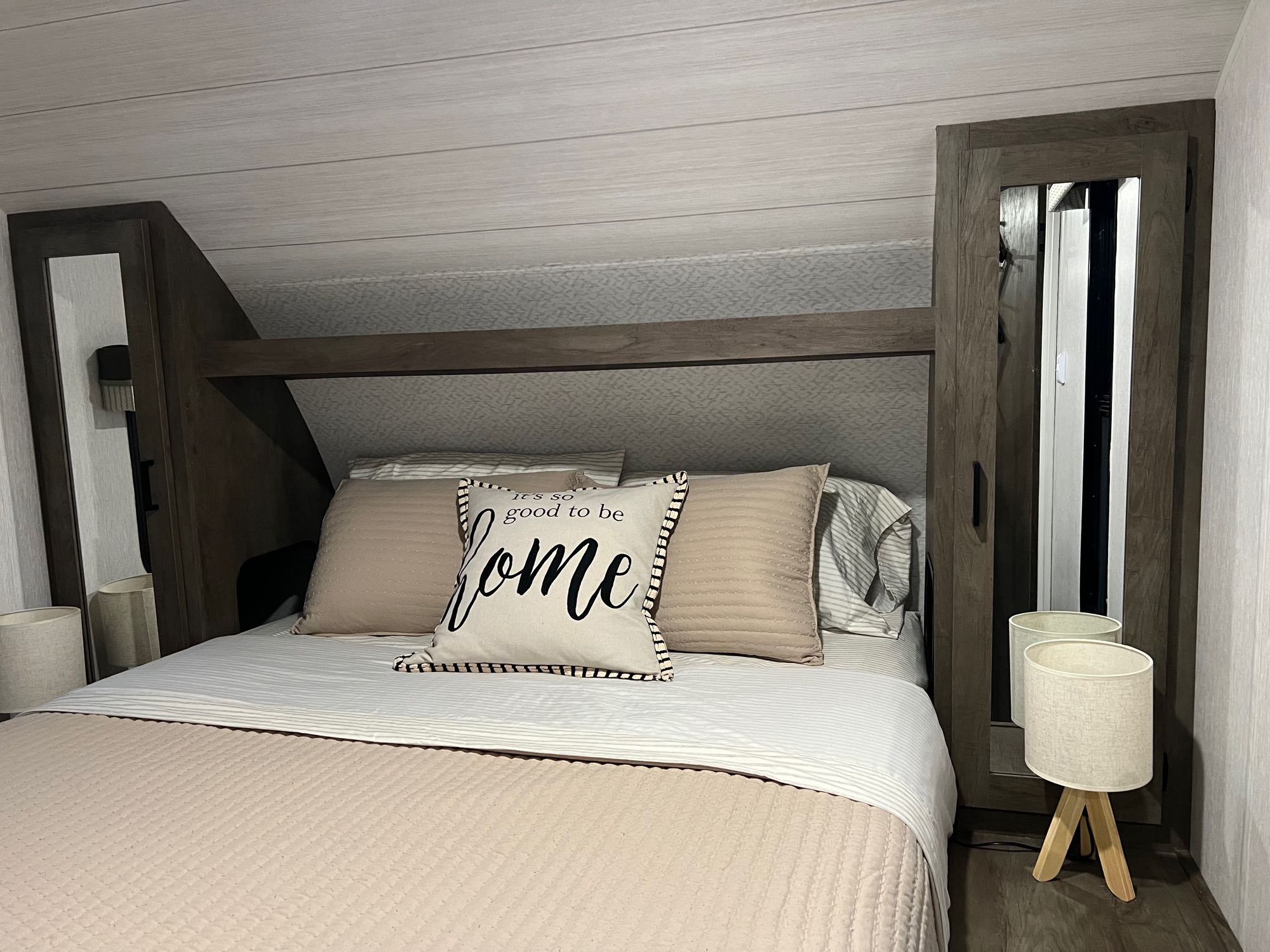 Comfortable RV rental bedroom featuring a cozy bed with beige and white bedding, built-in storage, and a bedside lamp.. Forest River Wildwood X-Lite 2023