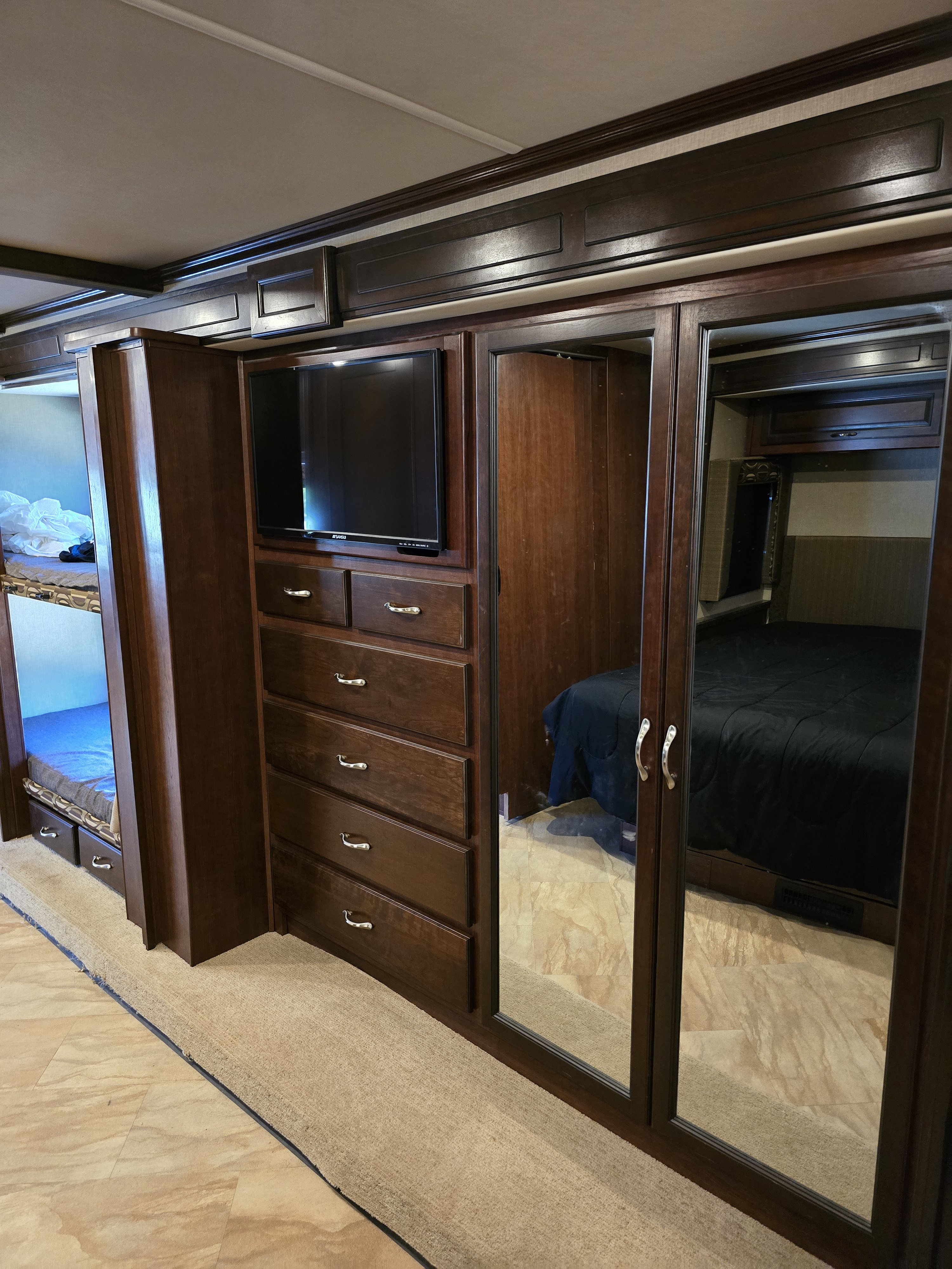 RV rental interior with dark wood cabinetry, featuring bunk beds, a TV, dresser, and mirrored closet reflecting a master bed.. Bounder Bounder Motorhome 2017