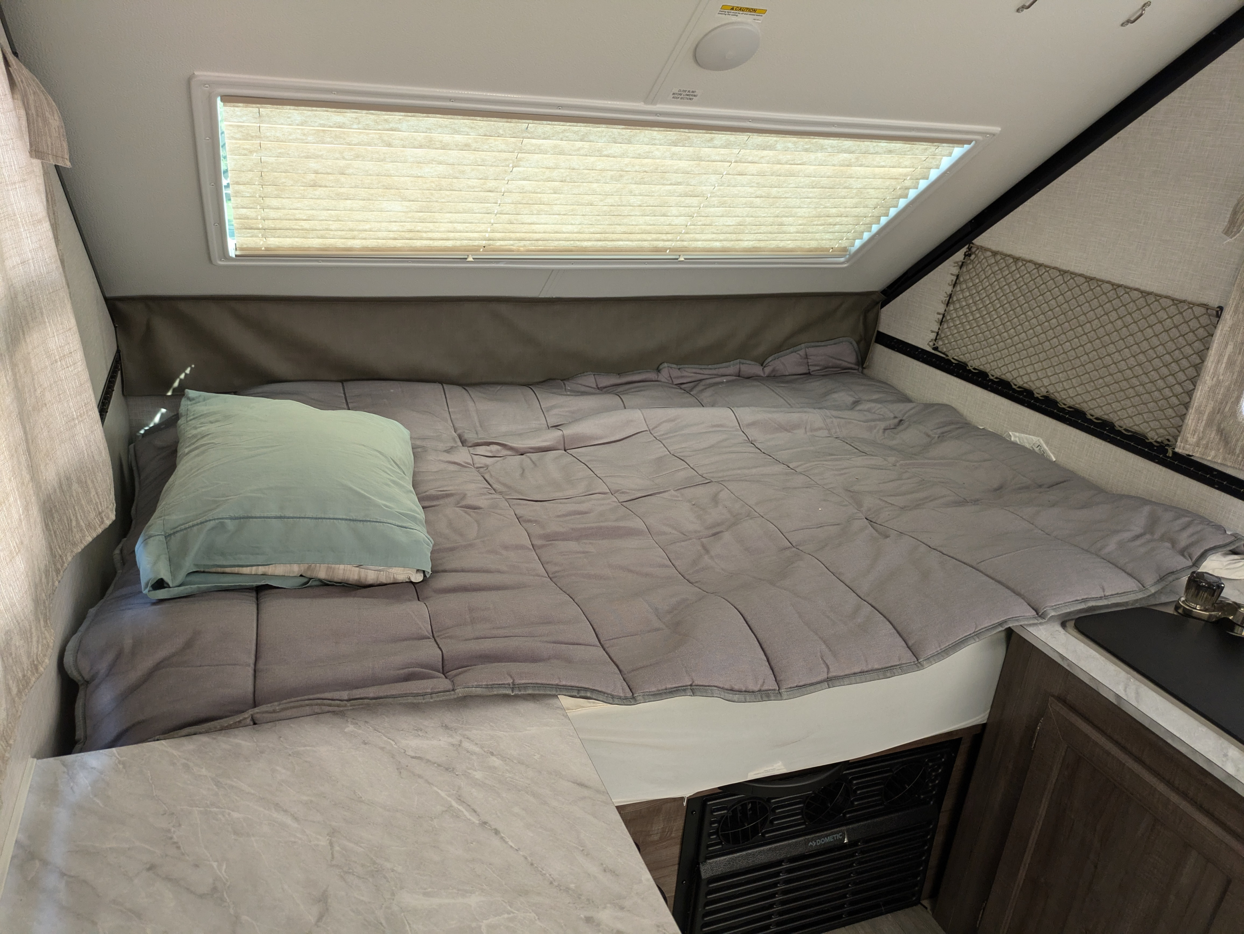 Cozy A-frame RV rental interior: bed, window with blinds, and compact living features. Ready for your next adventure!. Rockwood A122s 2023