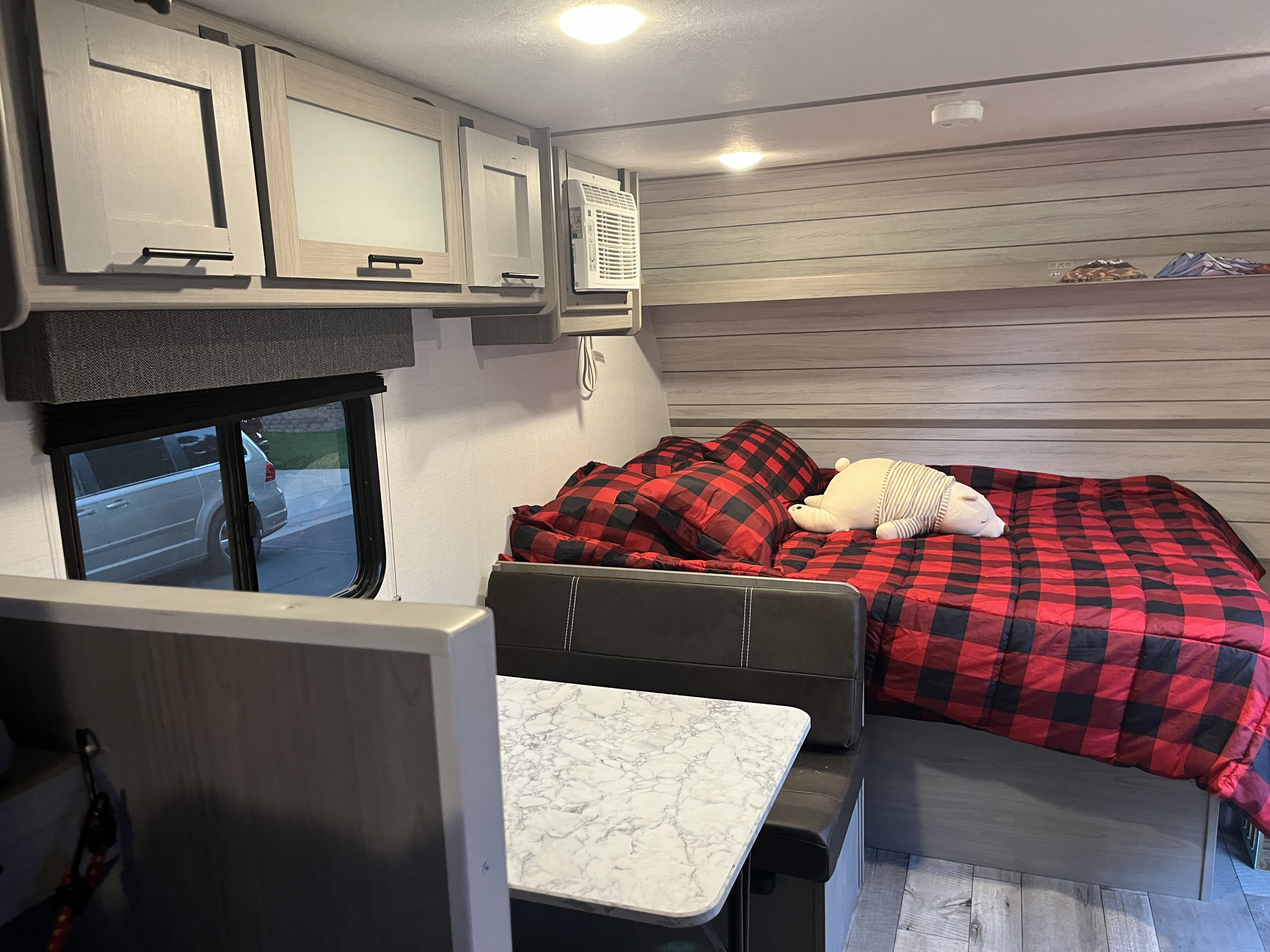 Cozy RV rental interior with red plaid bed, dining area, and overhead cabinets. Ideal for comfortable road trips.. COLEMAN LANTERN LT 17B 3990 2024
