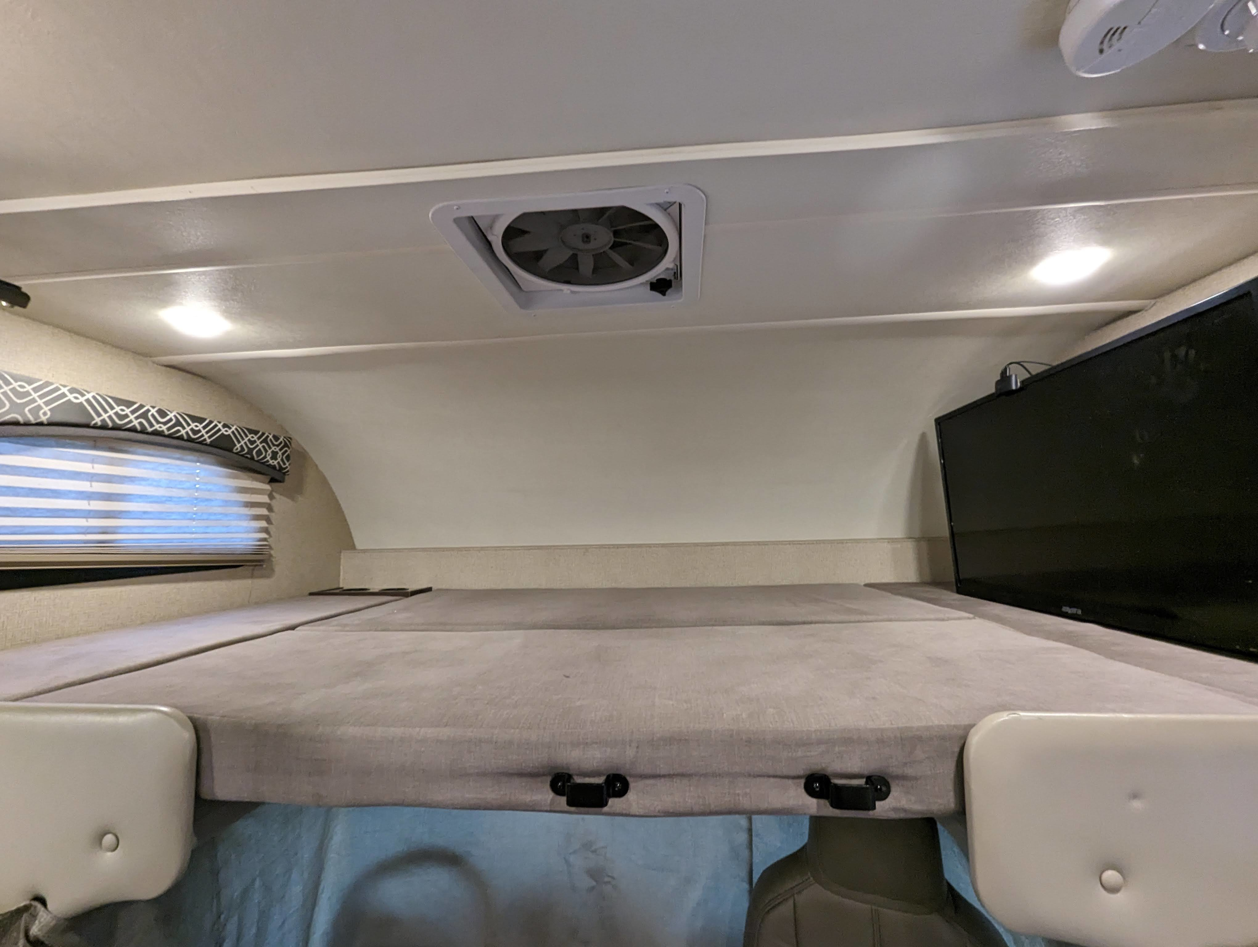 Cozy RV rental cab-over bunk with TV, fan, and window blinds, offering a comfortable sleeping area inside a recreational vehicle.. Thor Chateau 2019