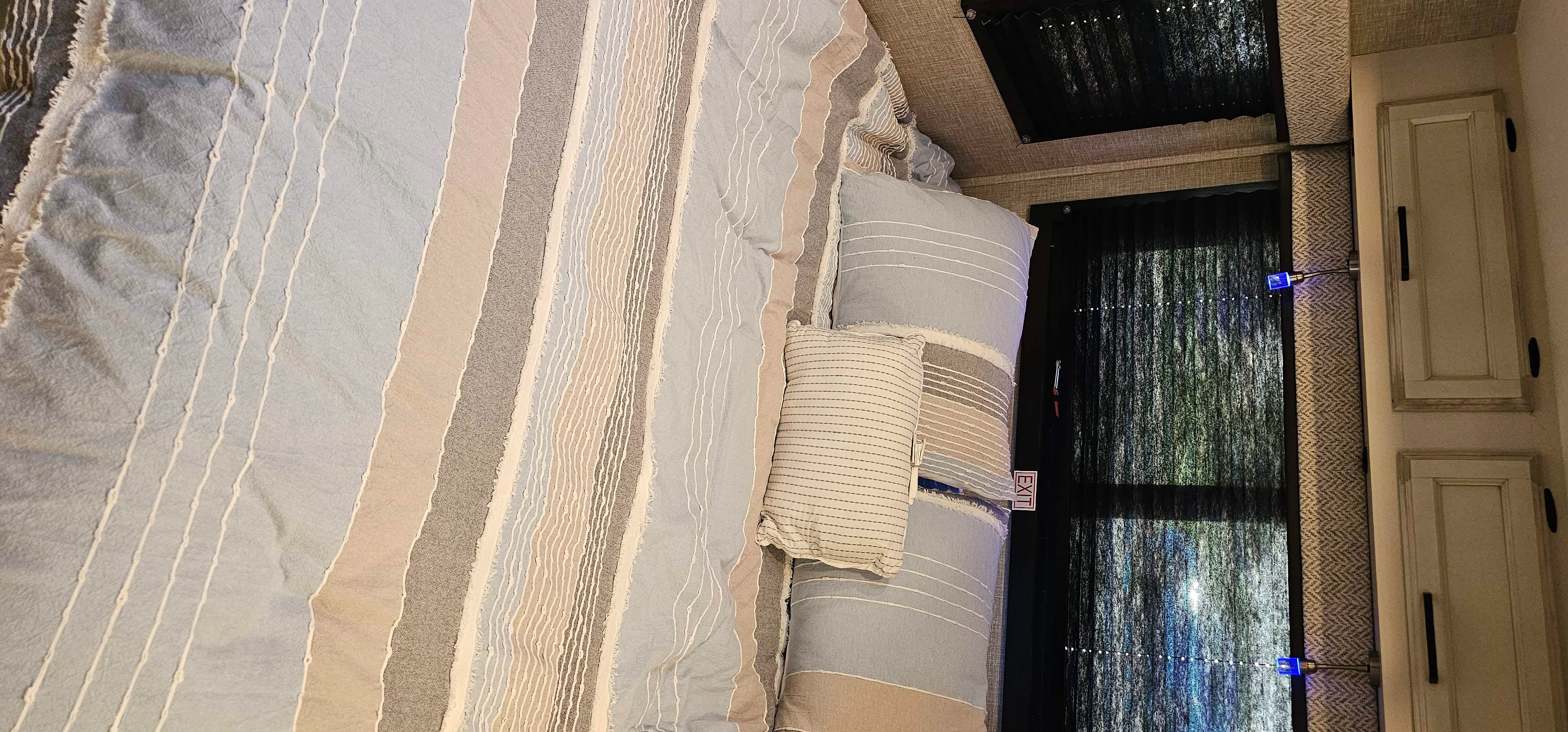 This cozy RV rental bedroom features a bed with striped blue, beige, and gray bedding, soft pillows, and dark window blinds.. Jayco Redhawk 2023