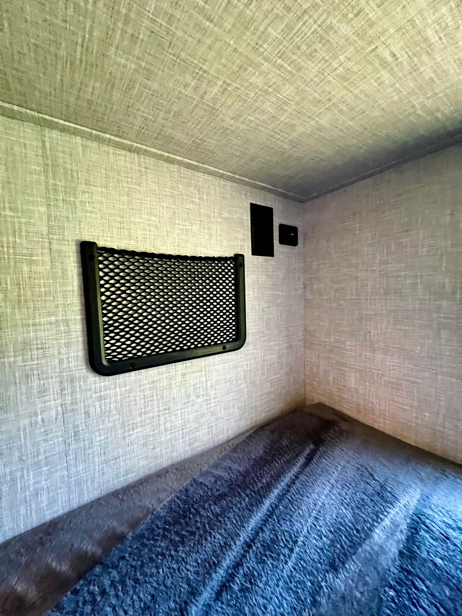 Inside an RV rental: a cozy bunk with textured walls, blue bedding, and a black mesh storage net.. Jayco Jay Feather 2024