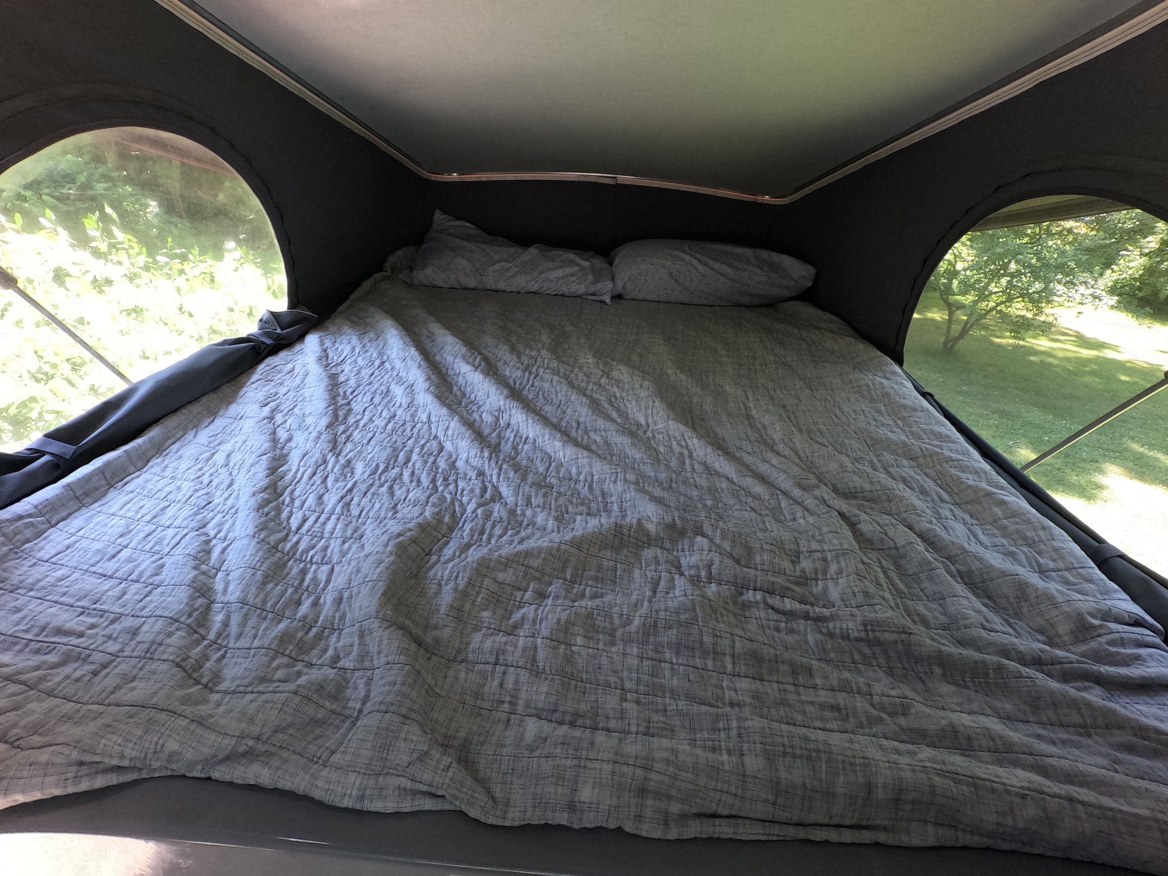 Inside an RV rental pop-top, a comfortable bed with grey bedding and pillows offers views of nature.. Winnebago Solis 2022