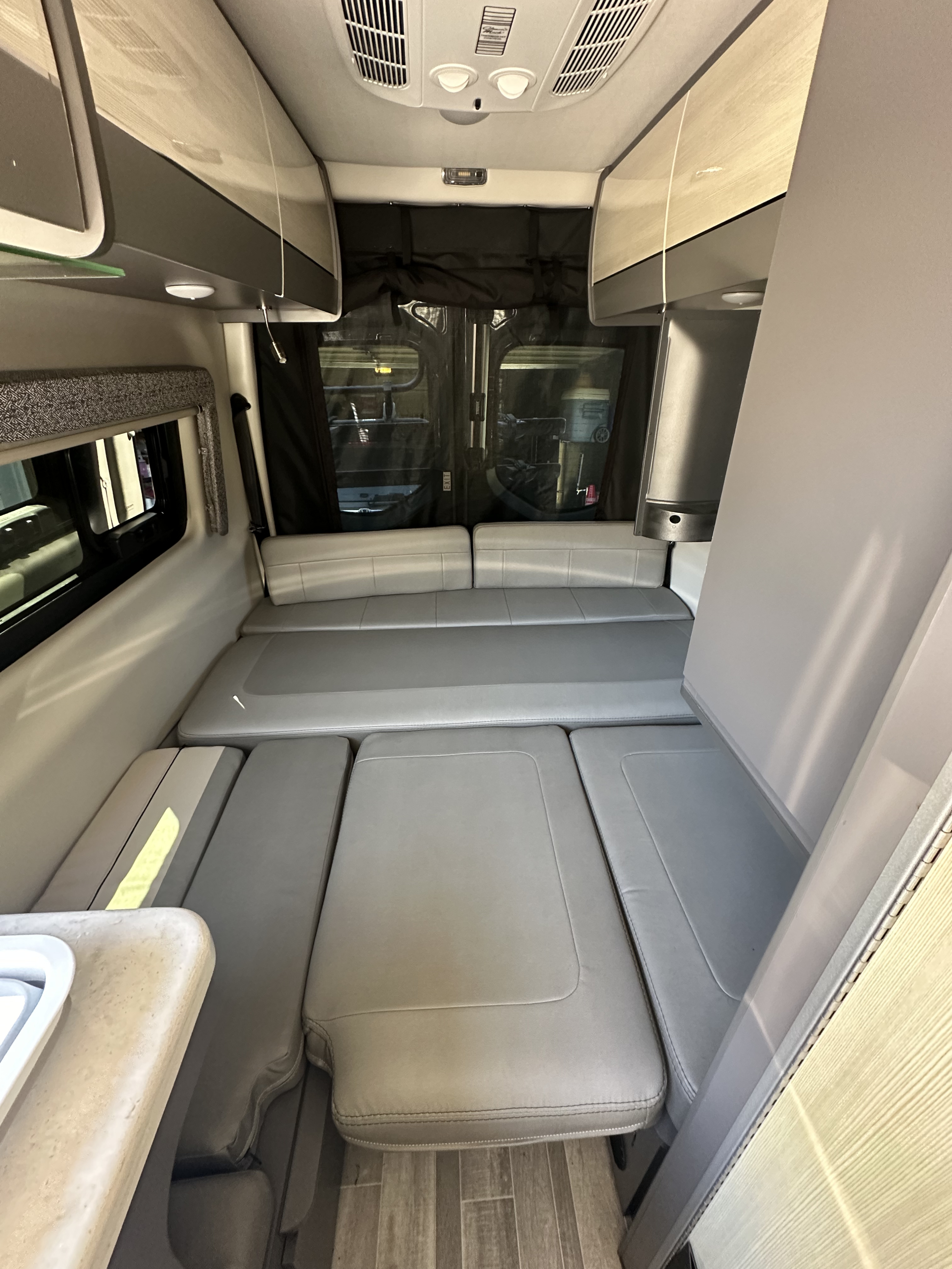 Interior view of a modern RV rental, featuring convertible light gray seating, overhead cabinets, and a side counter.. Thor Tranquility 2023