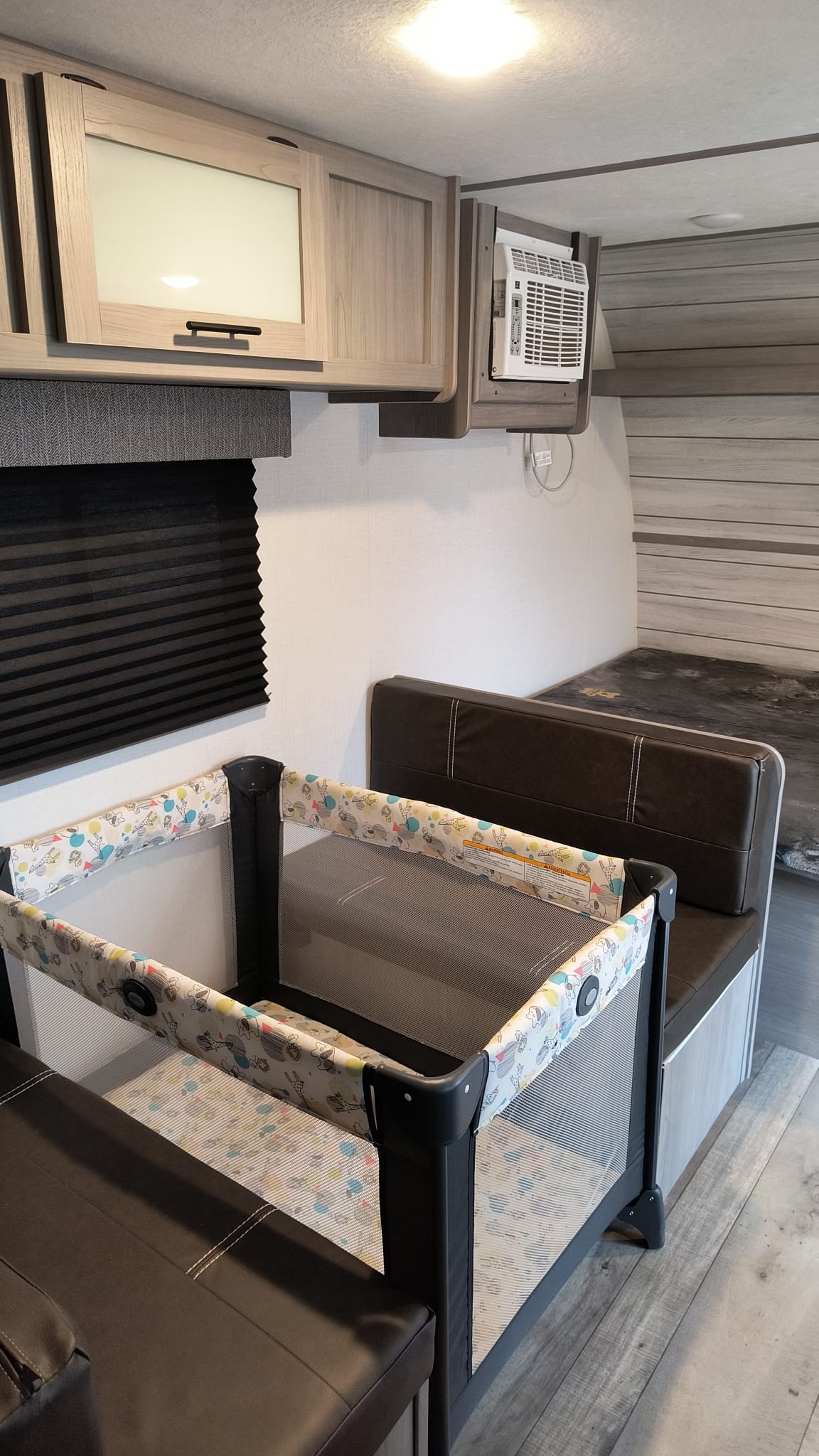 Inside an RV rental, a pack-n-play is set up between brown dinette benches. Overhead cabinets & AC unit visible.. Coachmen 17b 2024