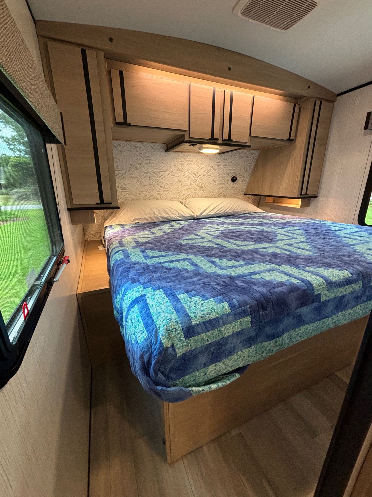 Cozy RV rental bedroom featuring a bed with a blue and green patterned quilt, overhead cabinets, and a window view.. Cruiser RV Radiance 2022