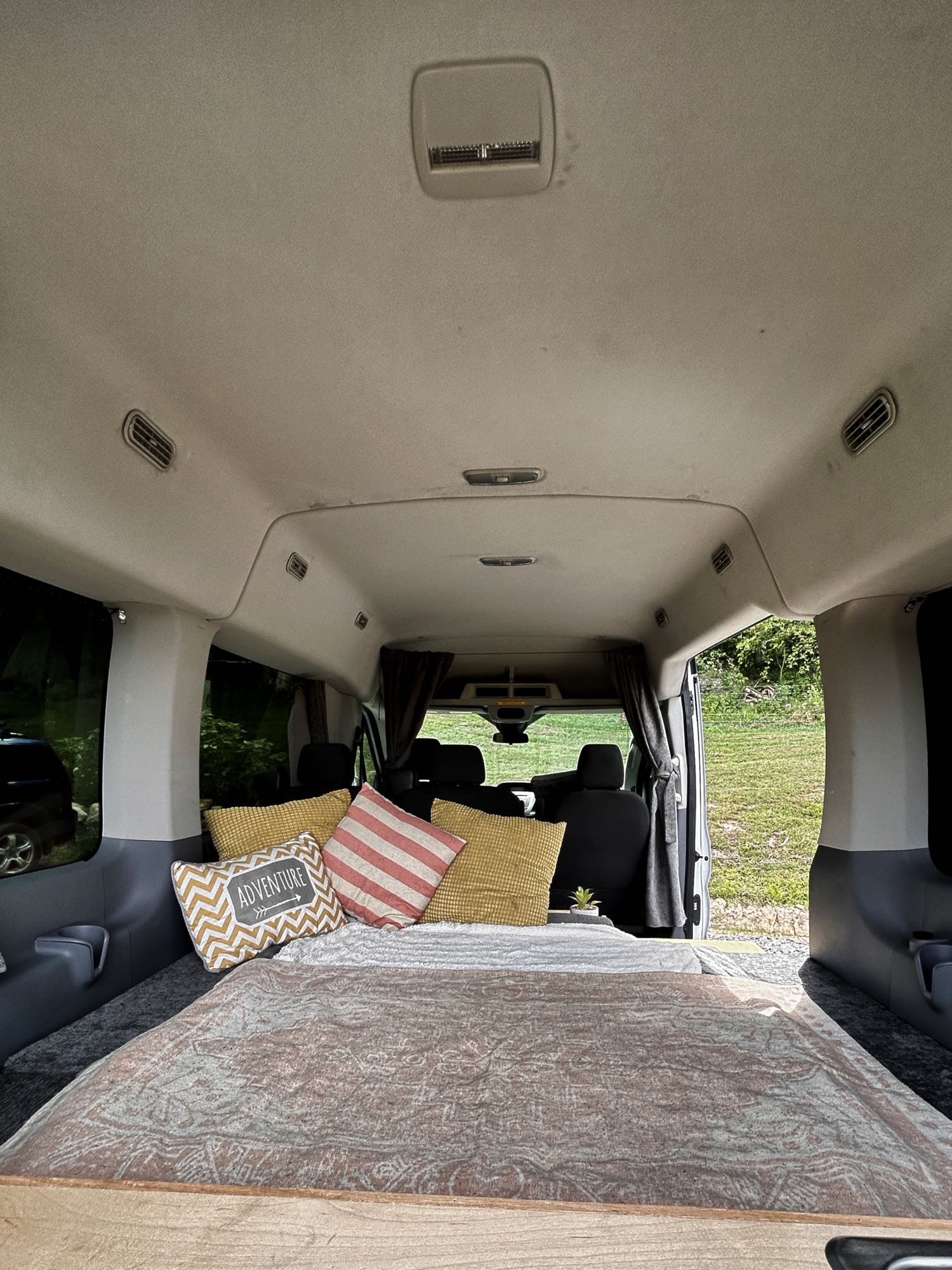Cozy RV rental interior featuring a bed with patterned blanket & "ADVENTURE" pillow, facing van cabin and nature view.. Ford Transit 150 XLT 2016