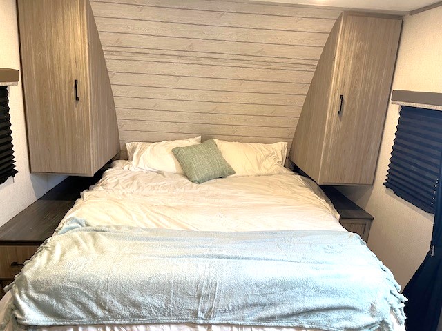 RV rental bedroom featuring a made bed with white bedding & blue blanket, flanked by nightstands & overhead storage.. Keystone Passport Sl series 2021