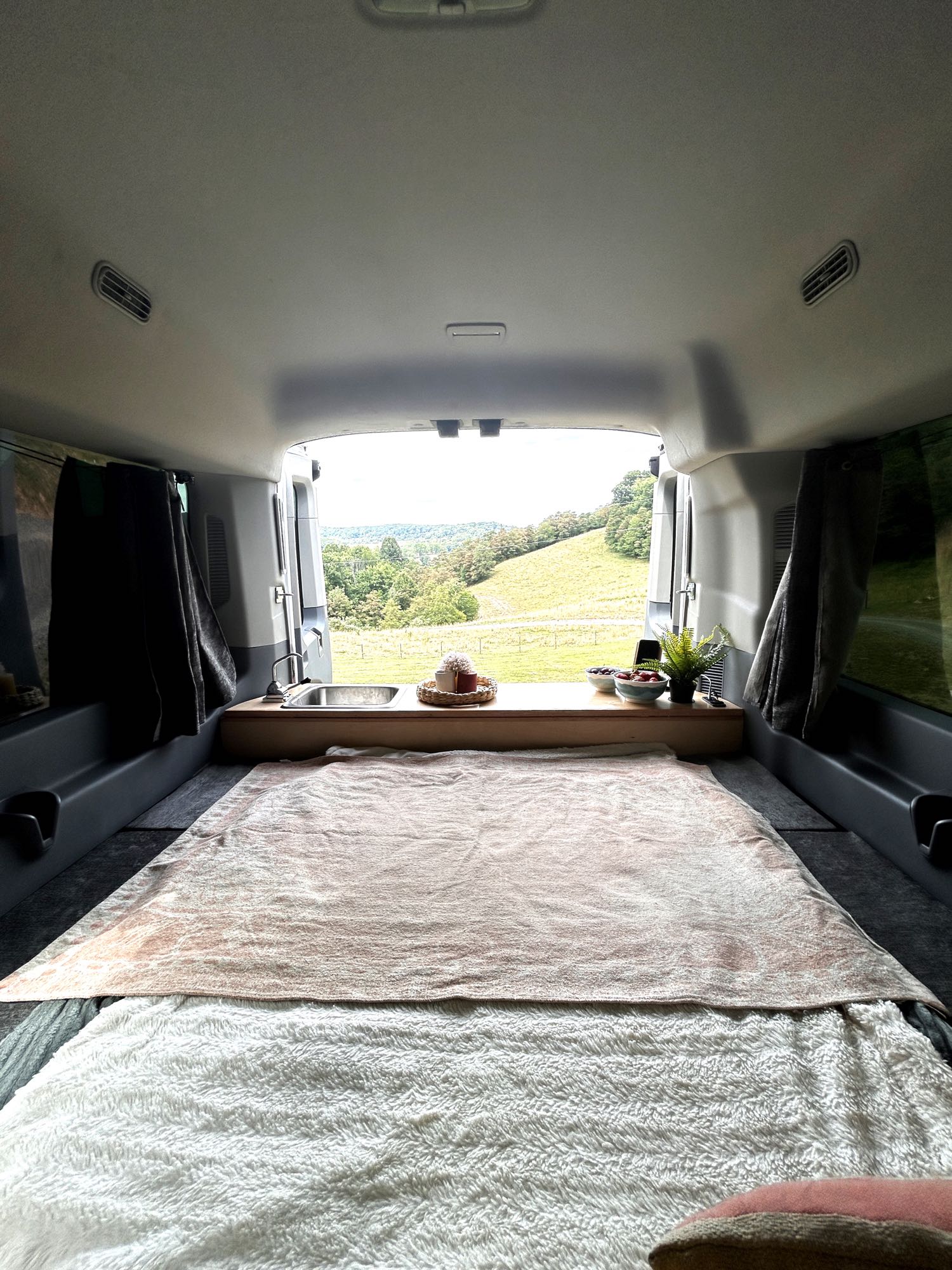Inside an rv rental: a cozy van interior with a bed, small kitchen counter, and a beautiful green hilly view from the back.. Ford Transit 150 XLT 2016