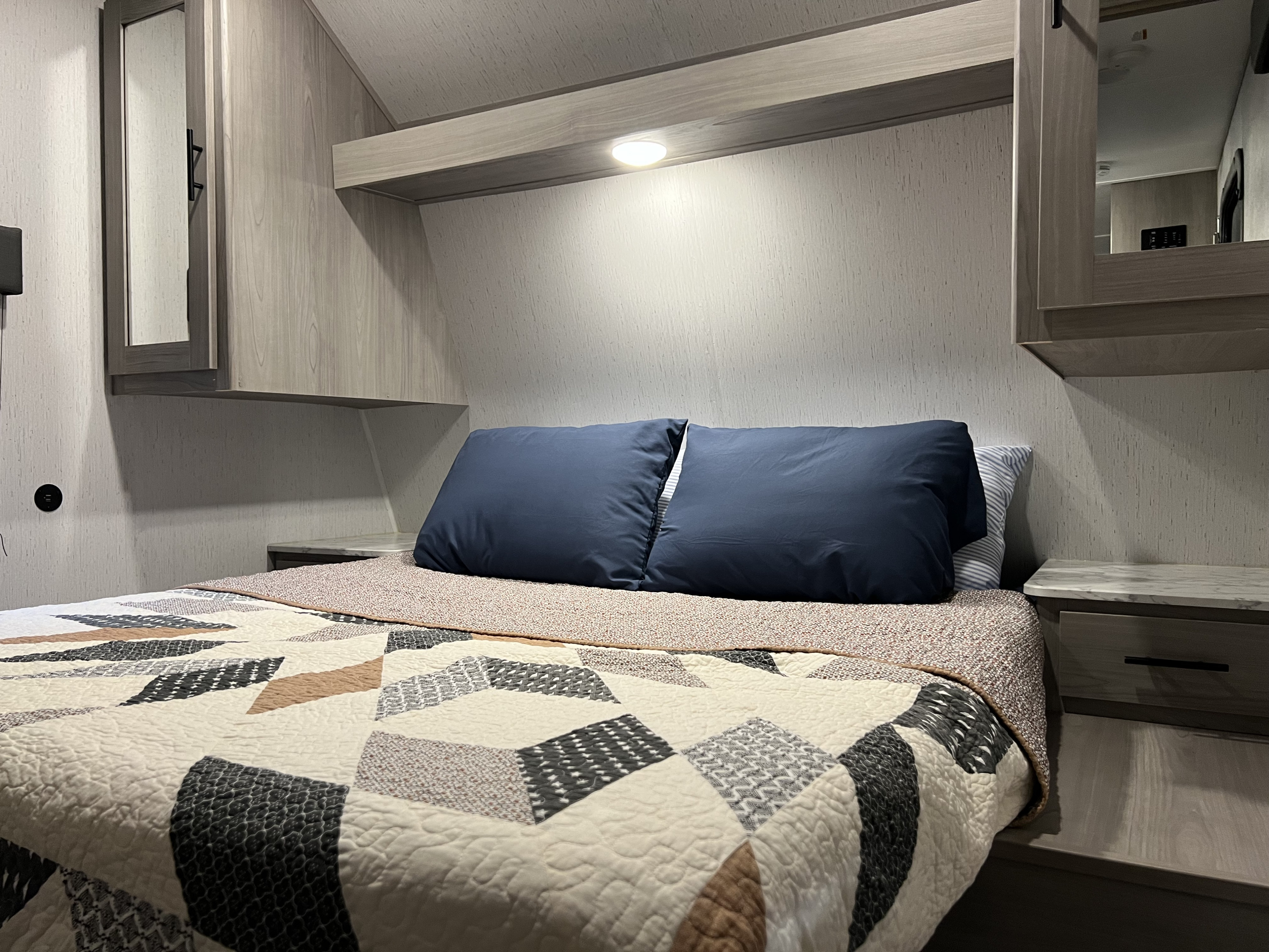 Cozy RV rental bedroom featuring a bed with a patterned quilt, blue pillows, built-in shelf light, and mirrored cabinet.. Dutchmen Coleman Lantern Toy Hauler 2022