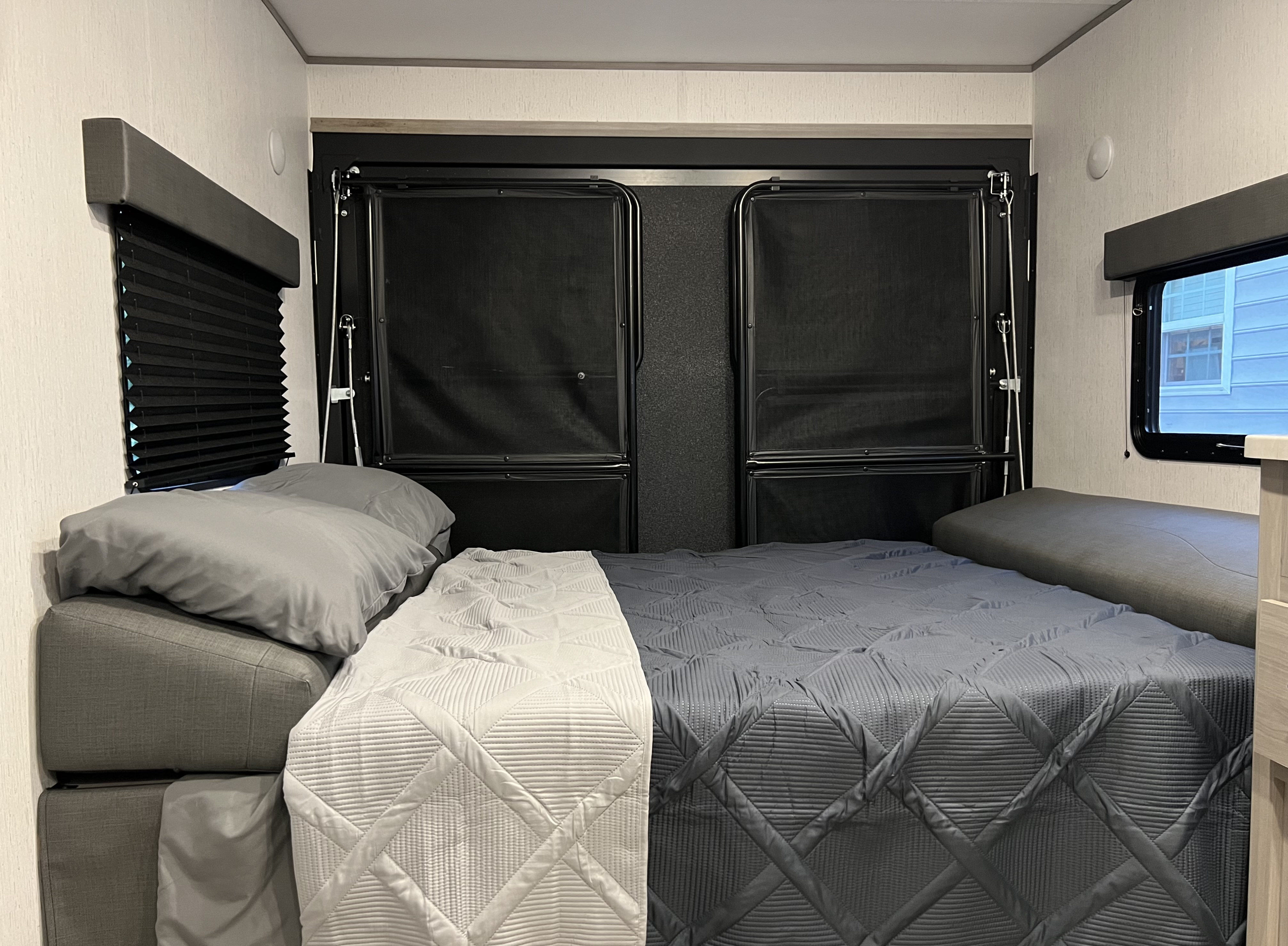 Inviting RV rental sleeping space with a neatly made grey bed, window with black blinds, and overhead mesh bunks.. Dutchmen Coleman Lantern Toy Hauler 2022