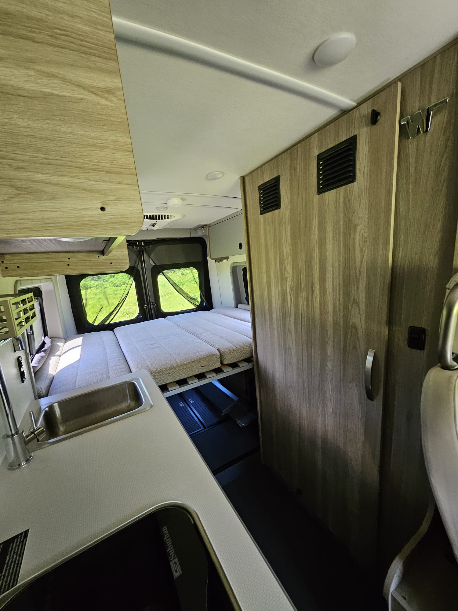 Modern RV rental interior: compact camper van features a convertible bed, kitchen sink, stove, and light wood cabinetry.. Winnebago Solis 2024