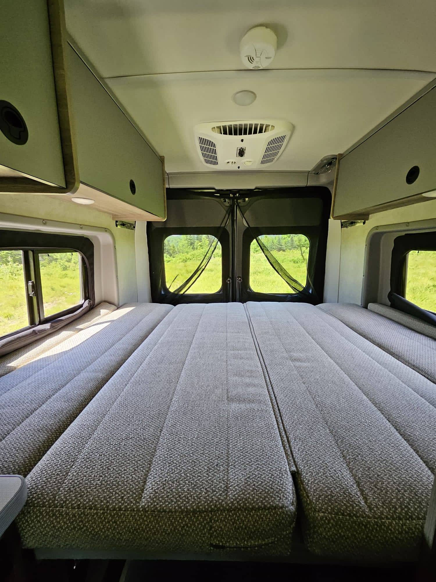 Inside a comfortable rv rental campervan, showing a large bed, overhead cabinets, and bright windows with views of green fields.. Winnebago Solis 2024