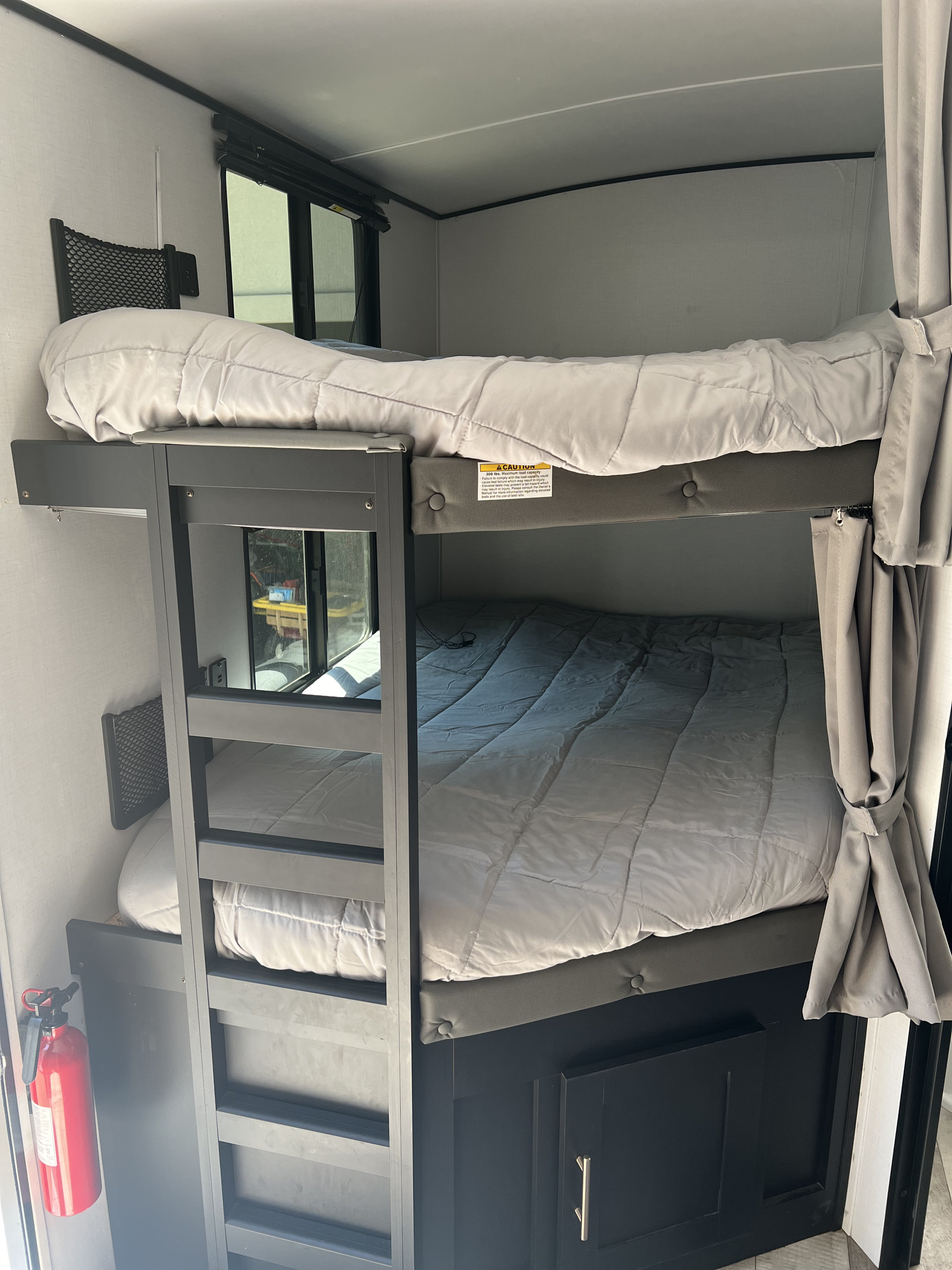 Interior view of an RV rental showing modern bunk beds with light gray bedding, windows, and a fire extinguisher for safety.. Keystone RV Springdale 2022