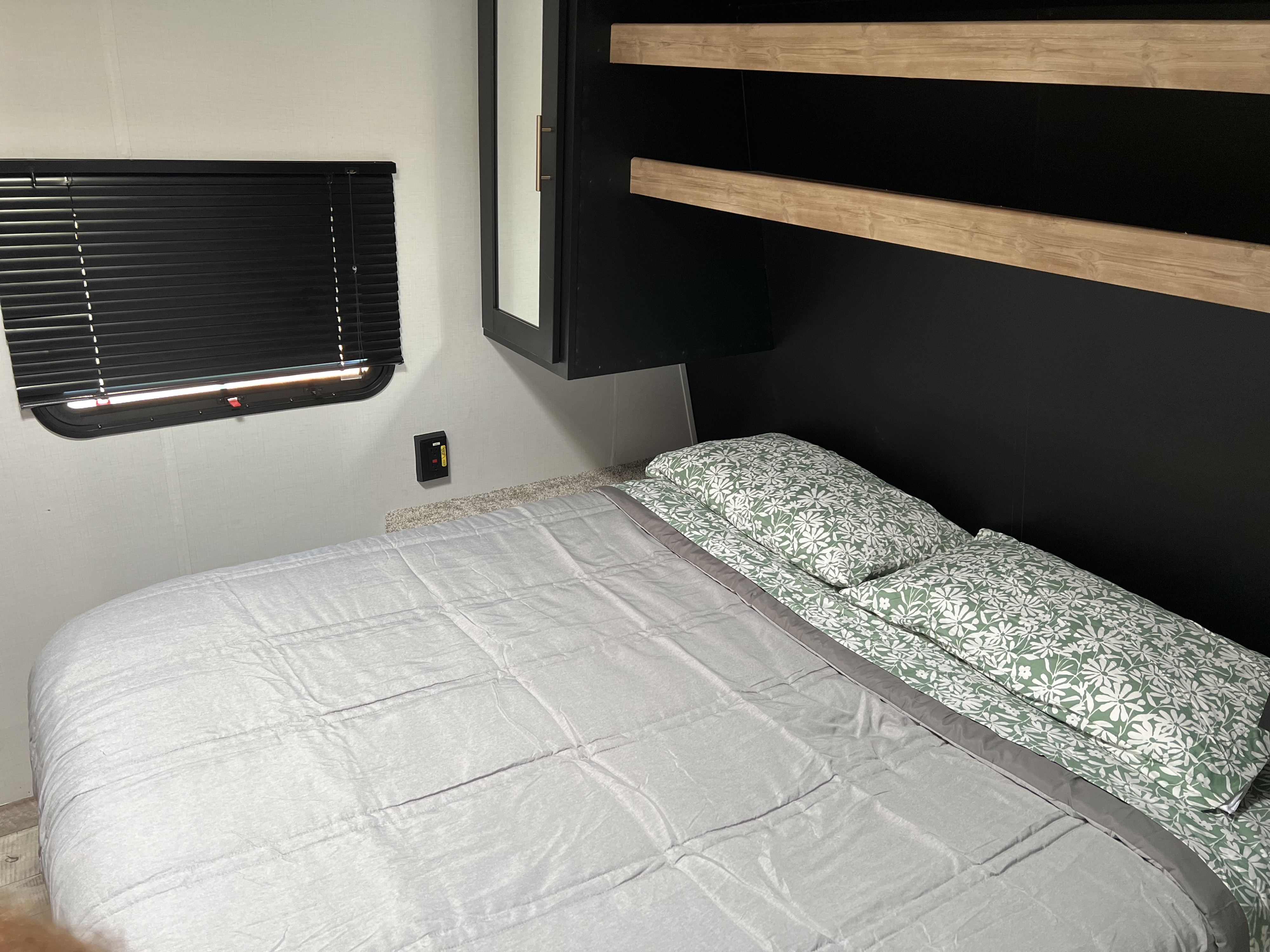 This RV rental bedroom features a bed with gray comforter & green floral pillows, black cabinets, wood shelves, and window.. Keystone RV Springdale 2022