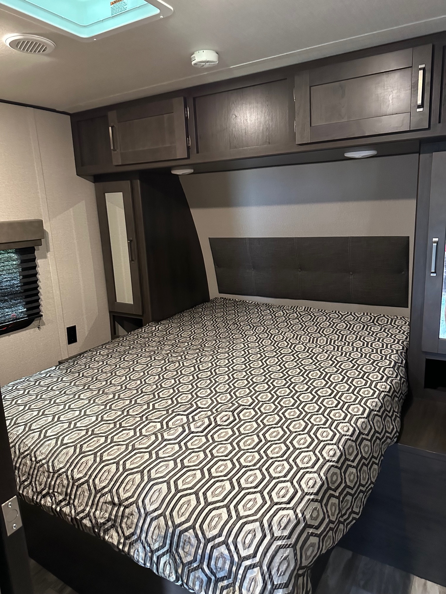 RV rental bedroom interior. Features a comfy bed with geometric comforter, overhead cabinets, and window for a cozy stay.. Grand Design Transcend 2020
