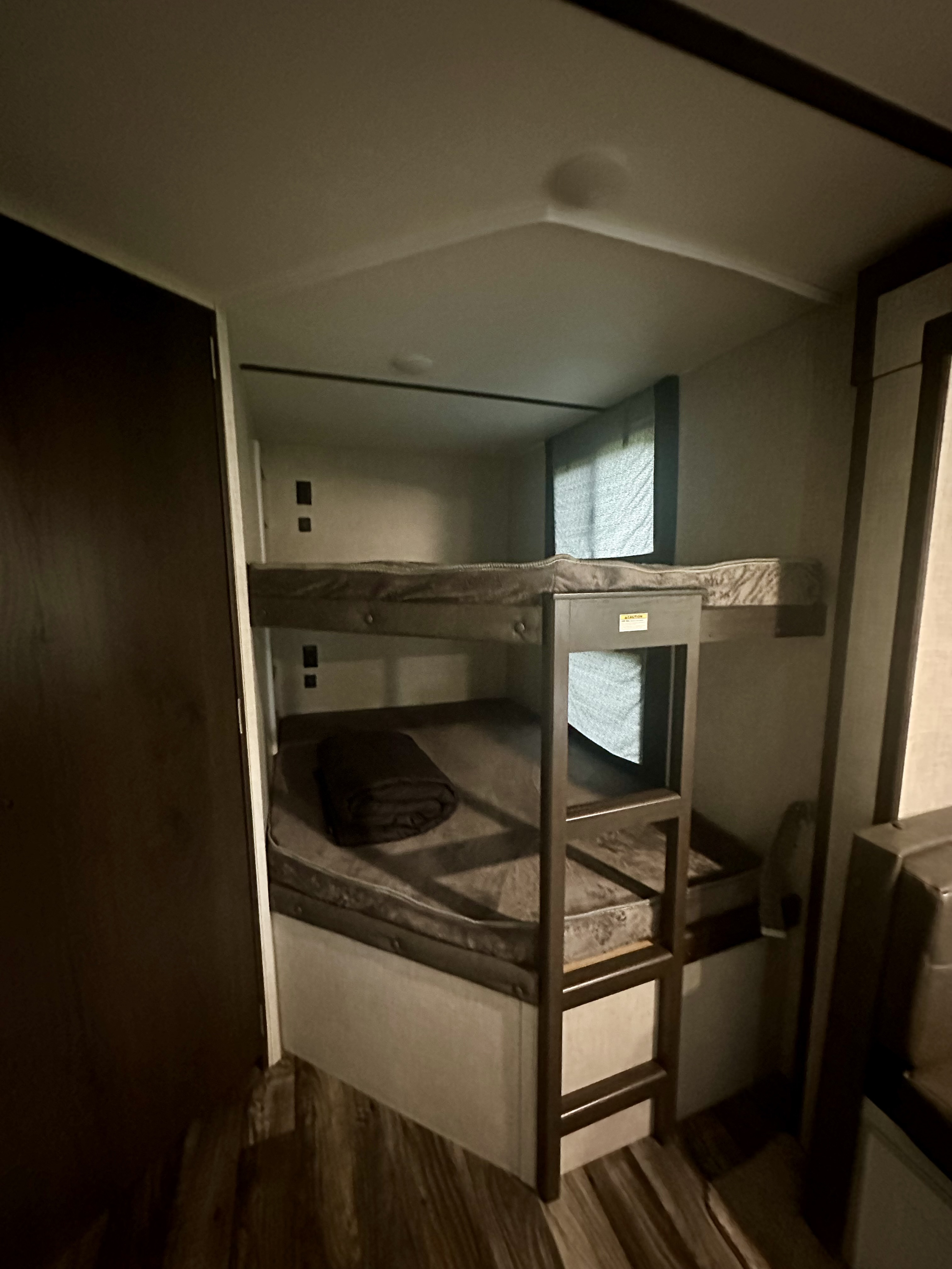 Interior view of an RV rental showing clean, grey bunk beds with a dark blanket on the lower bunk. Small window with shade.. Keystone RV Bullet Ultra Lite 2020