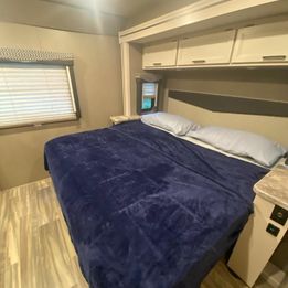 Comfortable RV rental bedroom featuring a large bed with dark blue blanket, window, and overhead cabinets.. Thor A.C.E. 2021