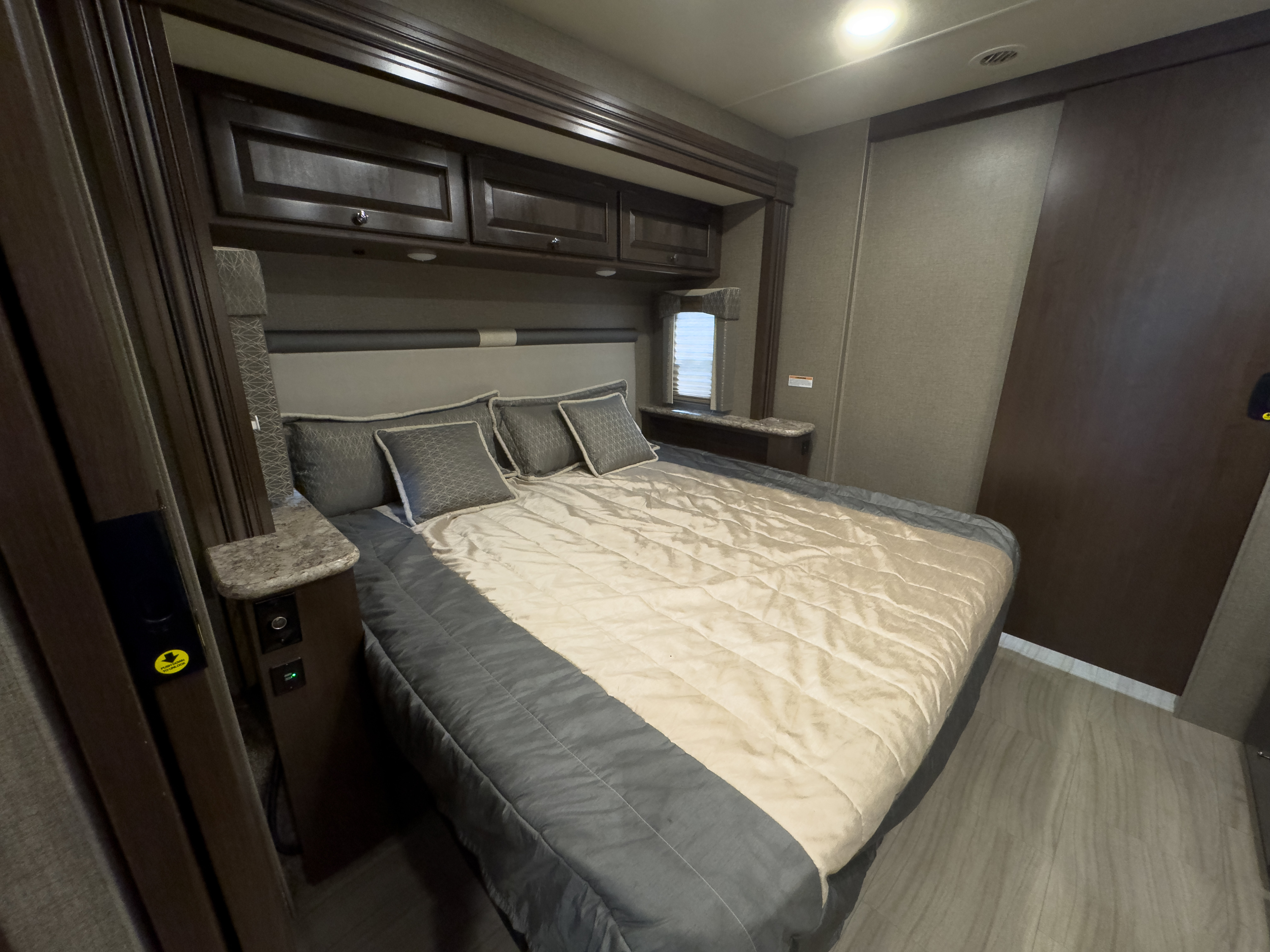 View inside an RV rental bedroom: queen bed with light & grey bedding, dark wood cabinets, and power outlets.. Thor Windsport 2020