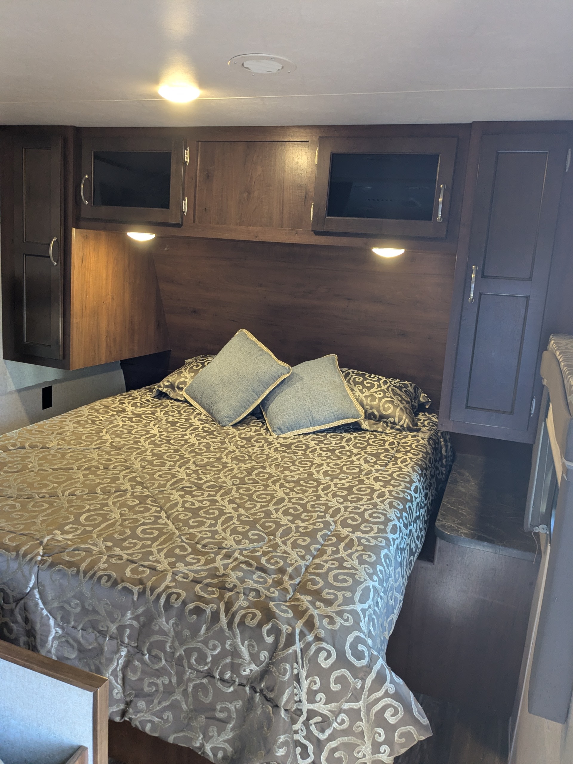 RV rental interior: A compact bedroom with a patterned bed, overhead cabinets, bedside outlet, and warm lighting.. Jayco Jay Flight 2017