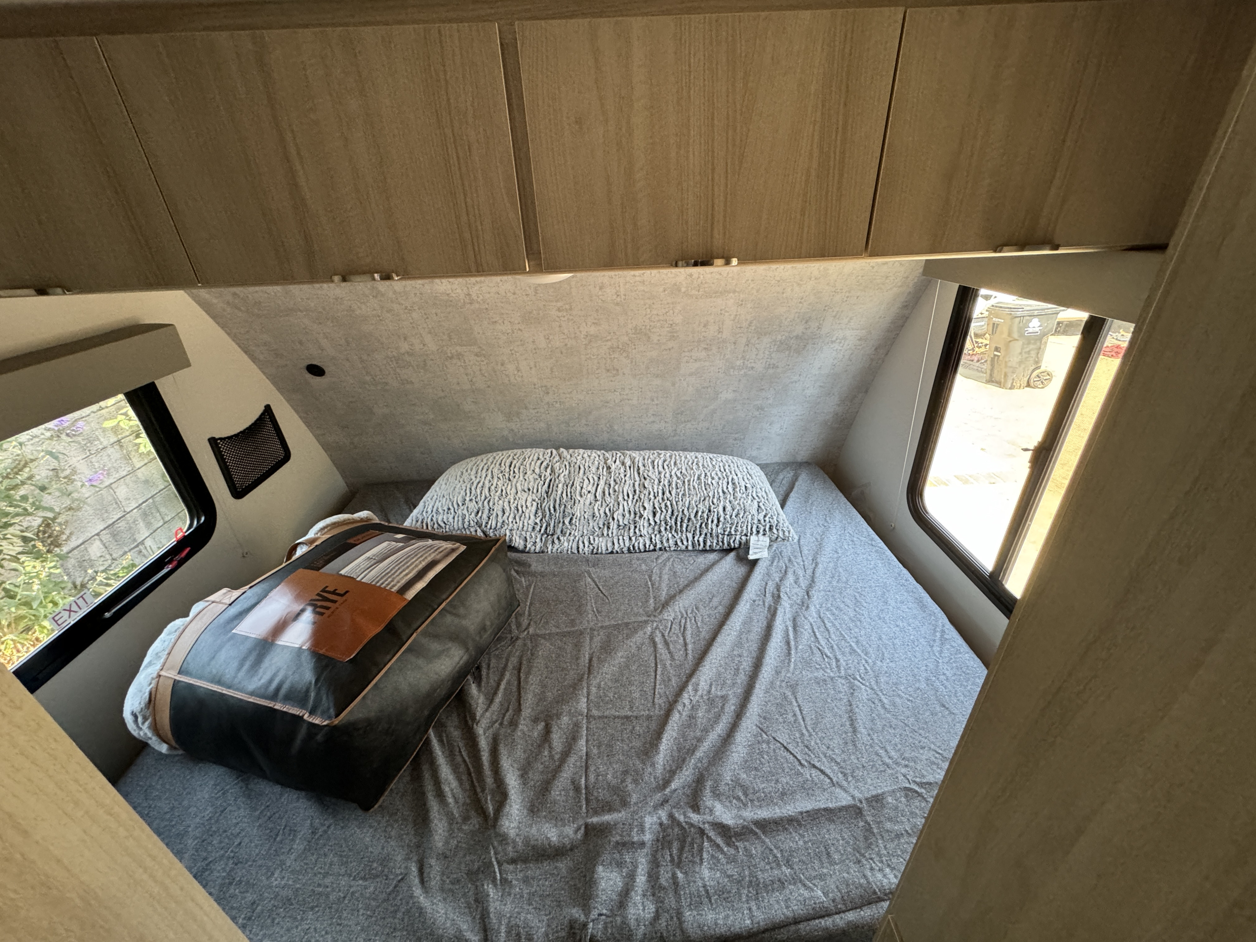 Inside a clean rv rental, showing a comfortable bed with grey sheets, overhead storage, and side windows in a compact camper.. Winnebago Micro Mini 2023