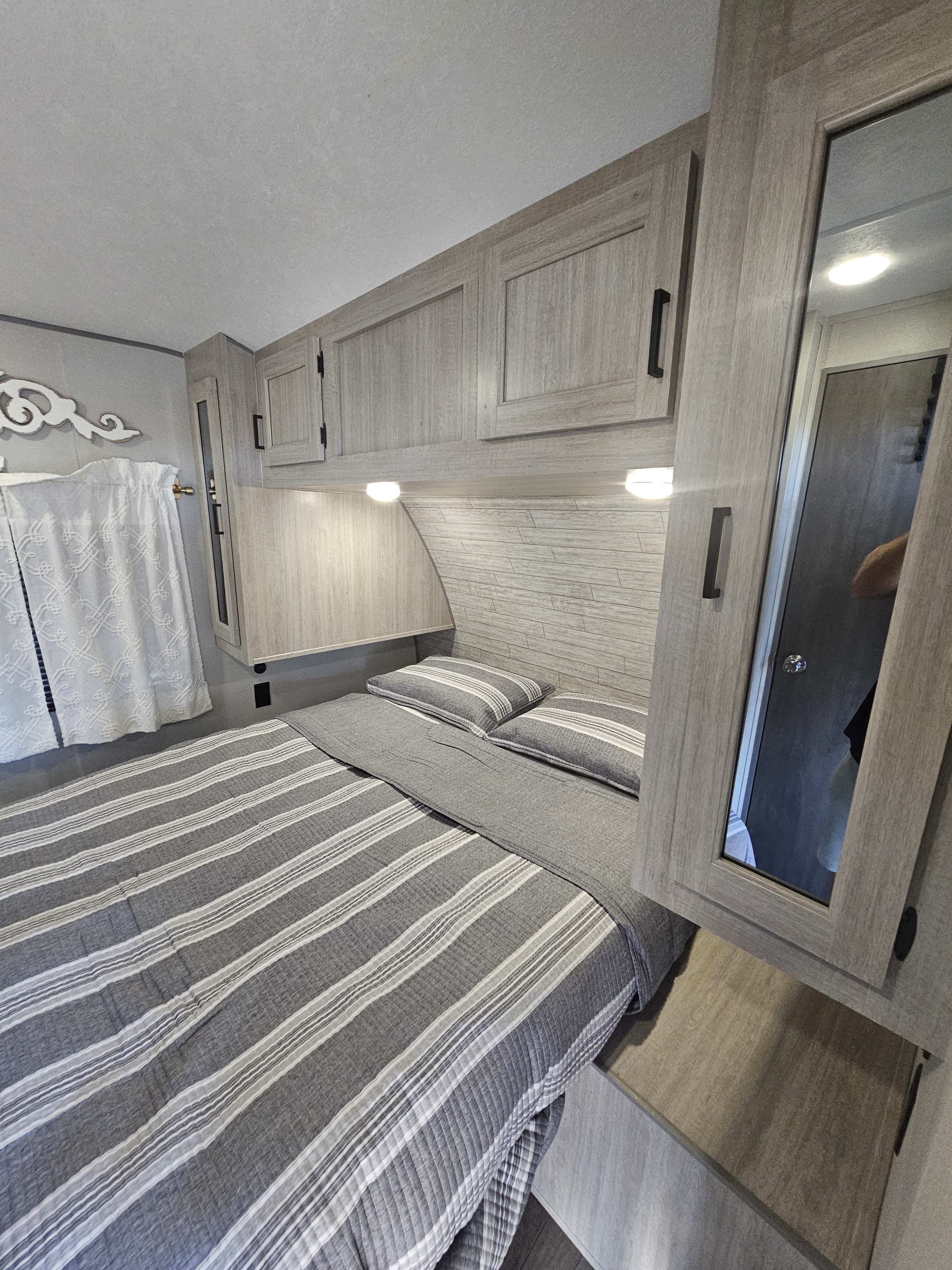 Light-toned RV rental bedroom with striped grey bedding, overhead cabinets, and subtle lighting.. Coachmen Catalina 2024