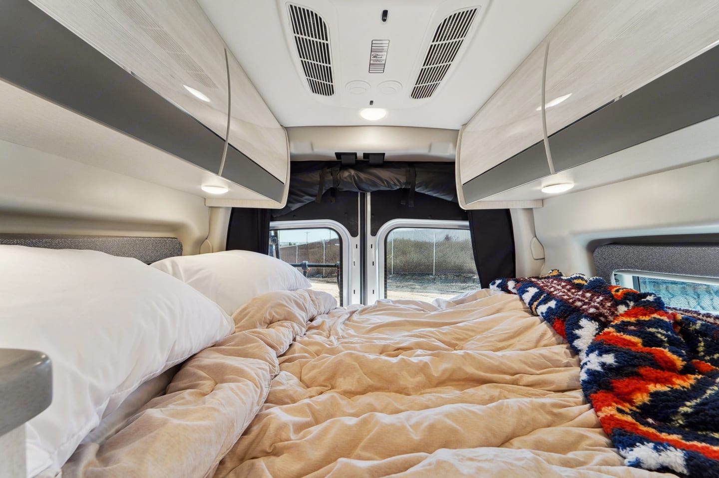 Cozy RV rental interior with a comfy bed, overhead storage, and rear windows showing the parked outdoor view.. Thor Motor Coach Sanctuary 2023