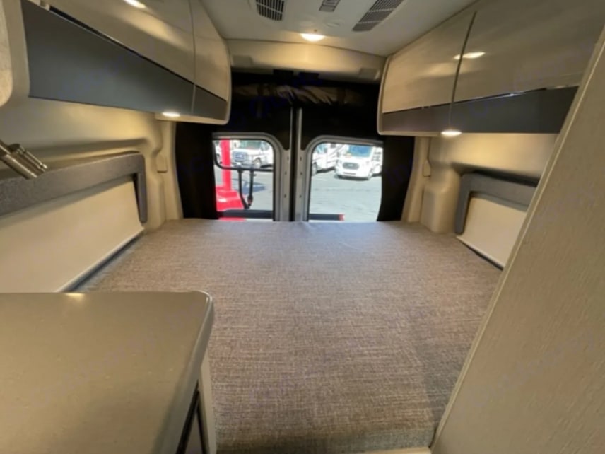 Modern RV rental interior: wide bed, overhead storage, and open rear doors revealing outside vehicles.. Thor Motor Coach Sanctuary 2023
