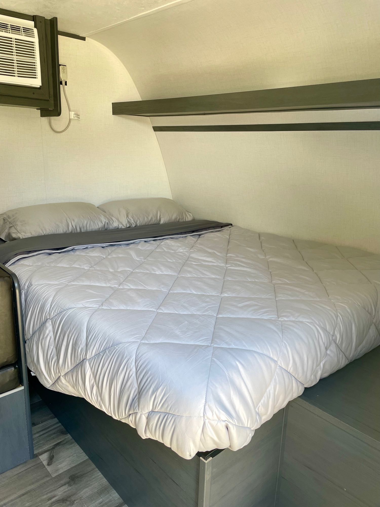 Inside a cozy RV rental: a comfortable bed with light grey bedding, wall-mounted AC unit, and built-in shelving.. Dutchmen Aspen Trail 2024