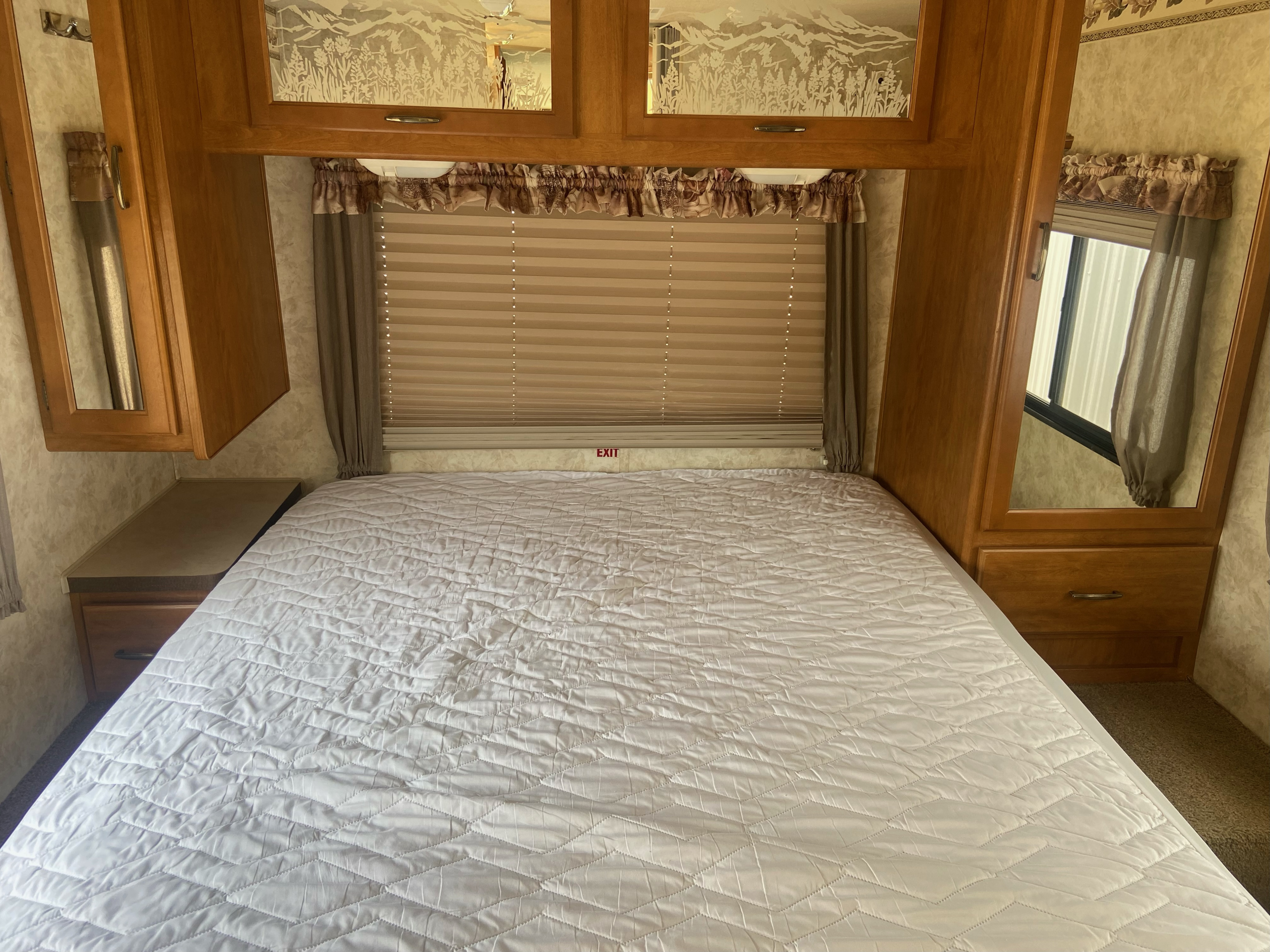 Cozy RV rental bedroom showing a large bed, overhead mirrored cabinets, and window with blinds. An exit sign is visible.. Fleetwood Terra 2003