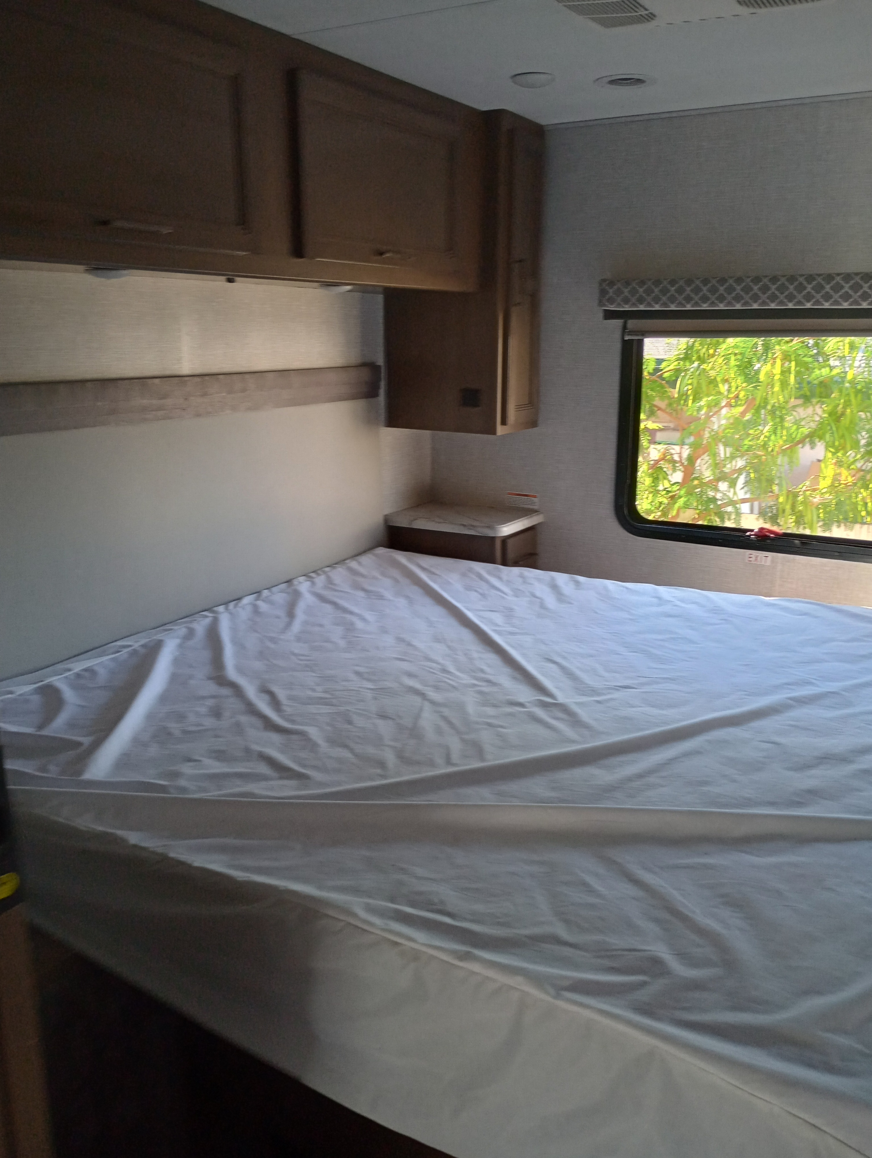 Cozy RV rental bedroom features a bed with white sheet, wood cabinets, nightstand, and window with foliage.. Thor Motor Coach Hurricane 2022