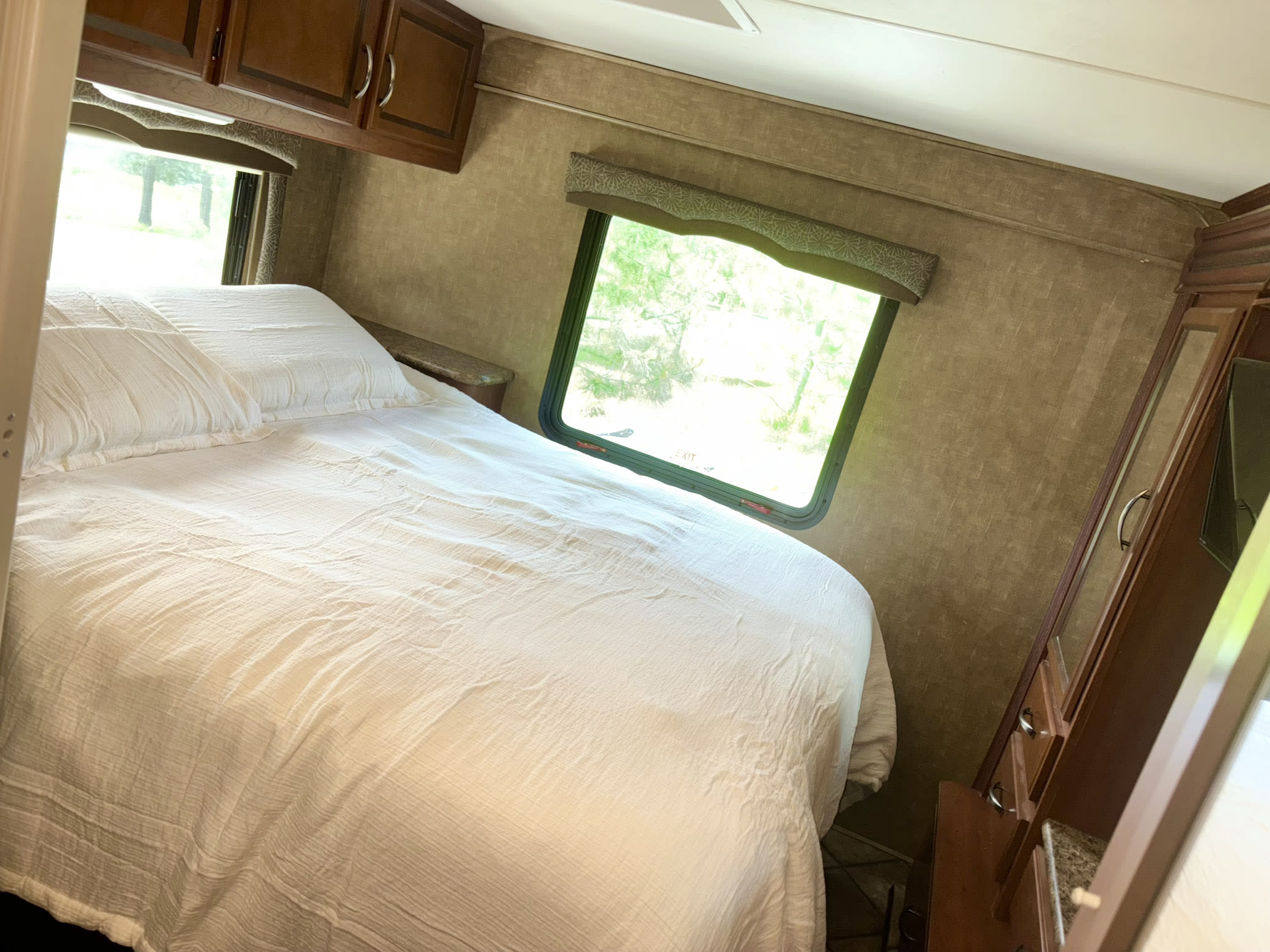 Spacious RV rental bedroom featuring a large white bed, overhead cabinets, and windows with a peaceful green view.. Thor Chateau 2014